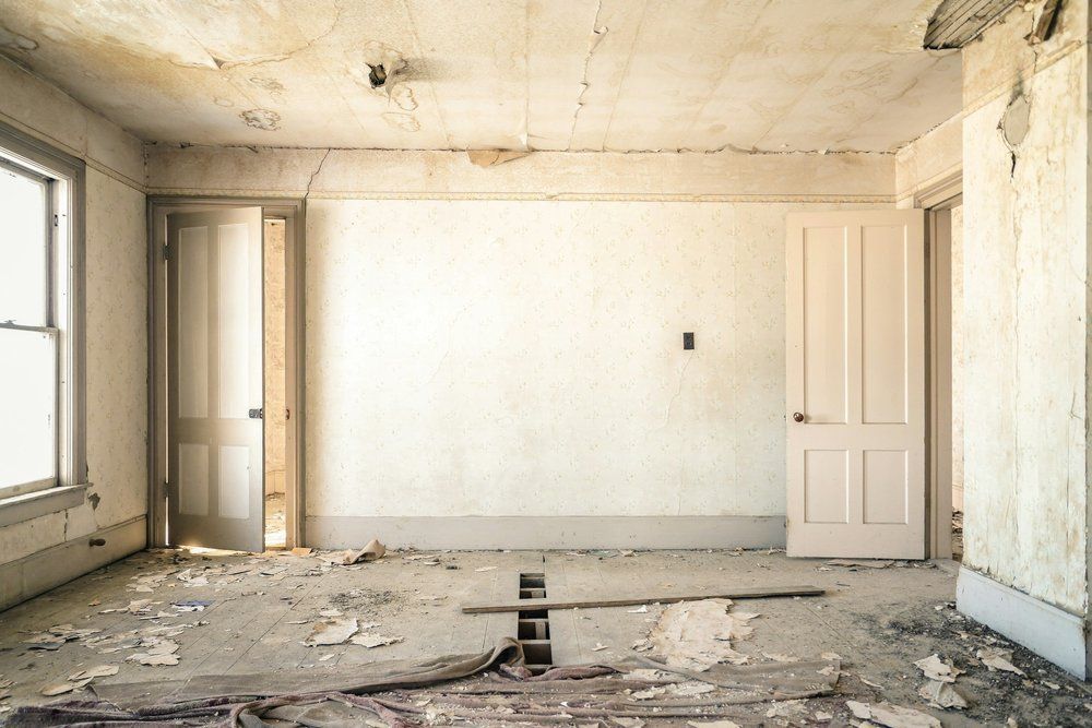 An empty, dilapidated room with peeling paint, open doors, and damaged floorboards.