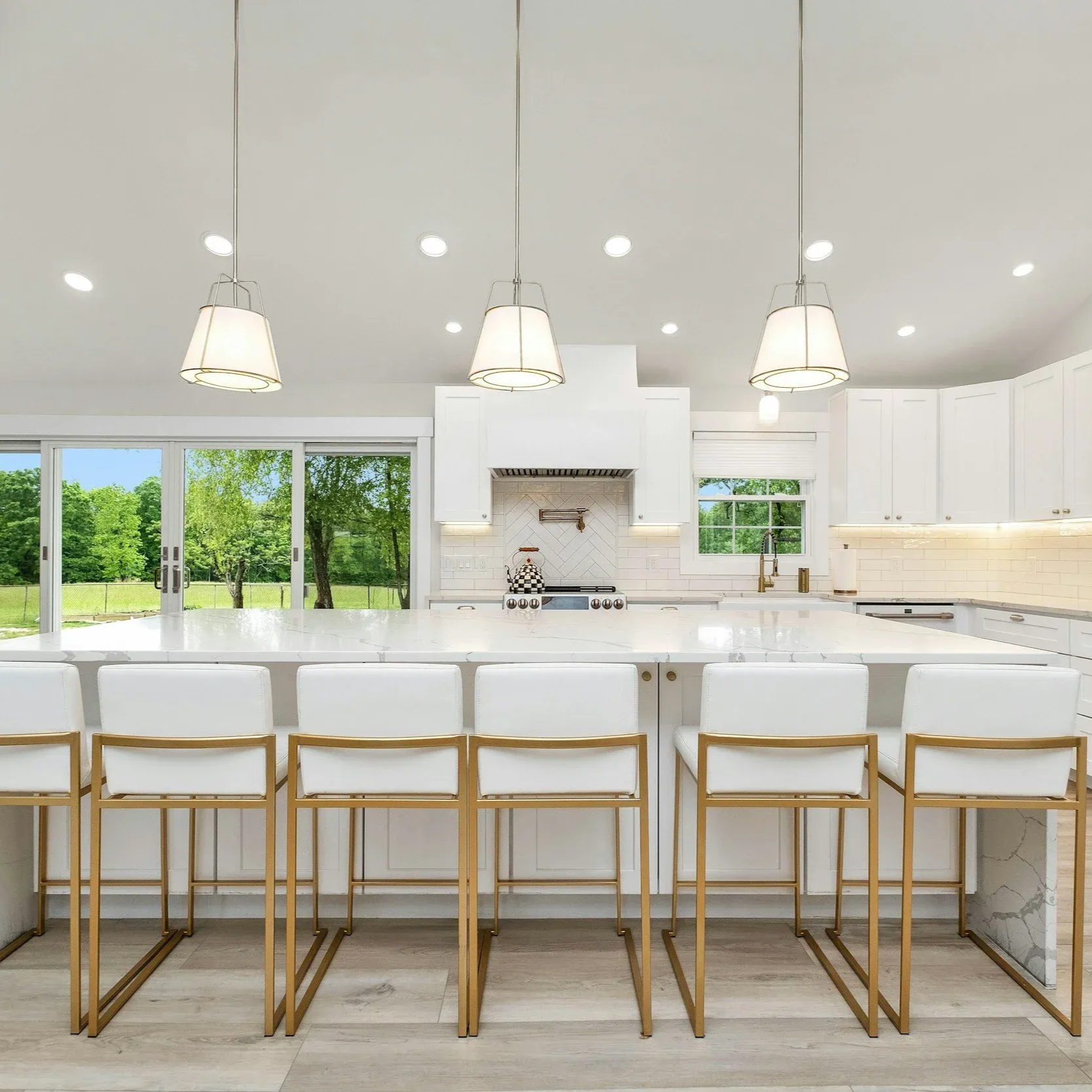 White kitchen with island, seating, pendant lights, and a view of green trees through a window.