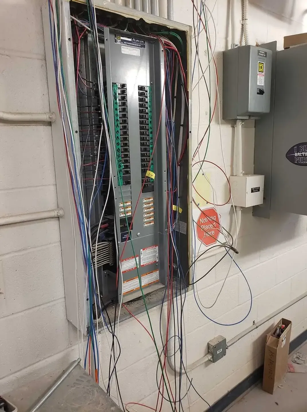 An open electrical breaker panel with exposed wires hanging down, mounted on a white cinder block wall.