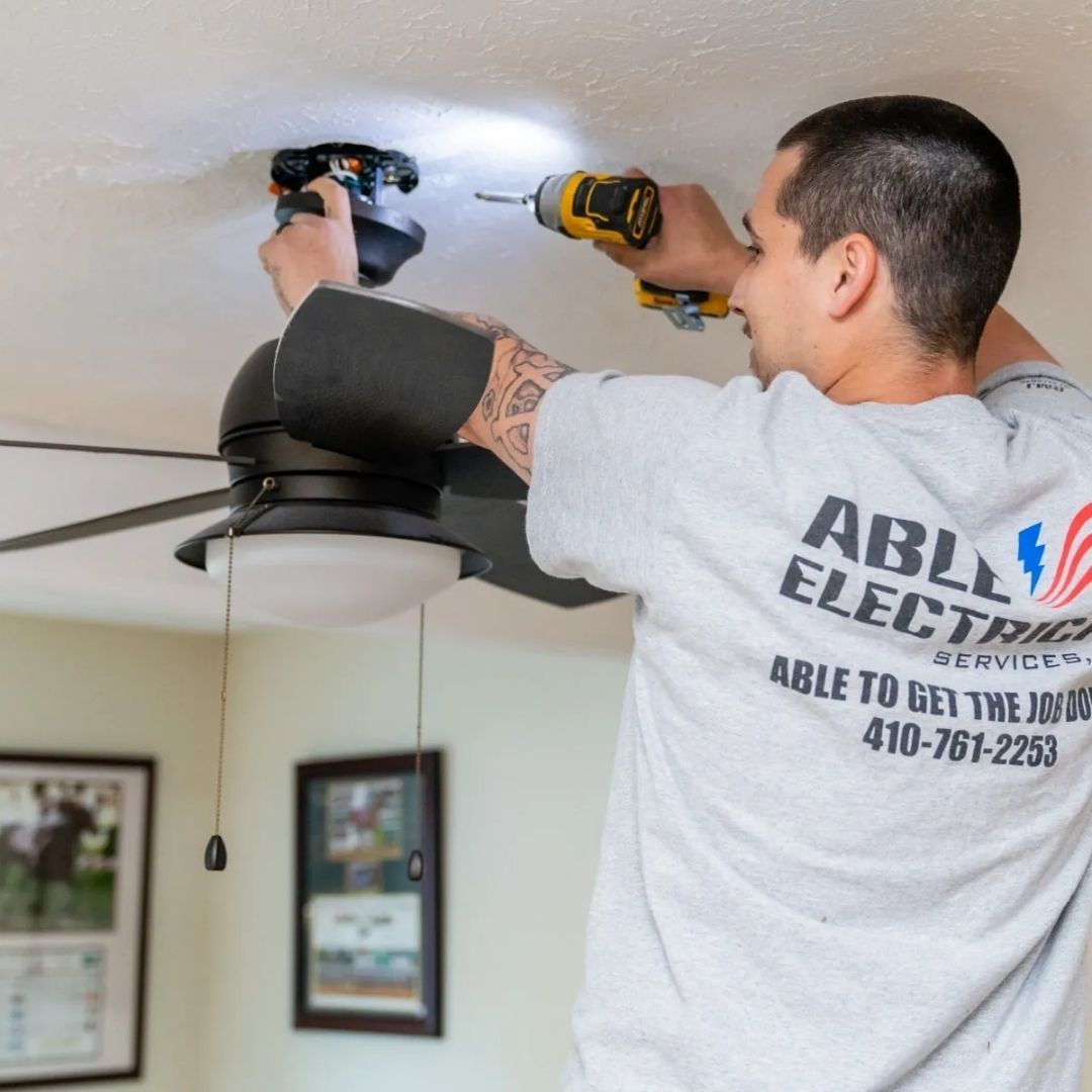 An electrician in an Able Electrical Services t-shirt drills a ceiling fan mount into a white ceiling.