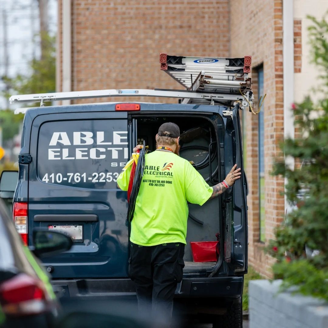 A technician in a neon yellow shirt loads equipment into an Able Electric van with a ladder rack on top.