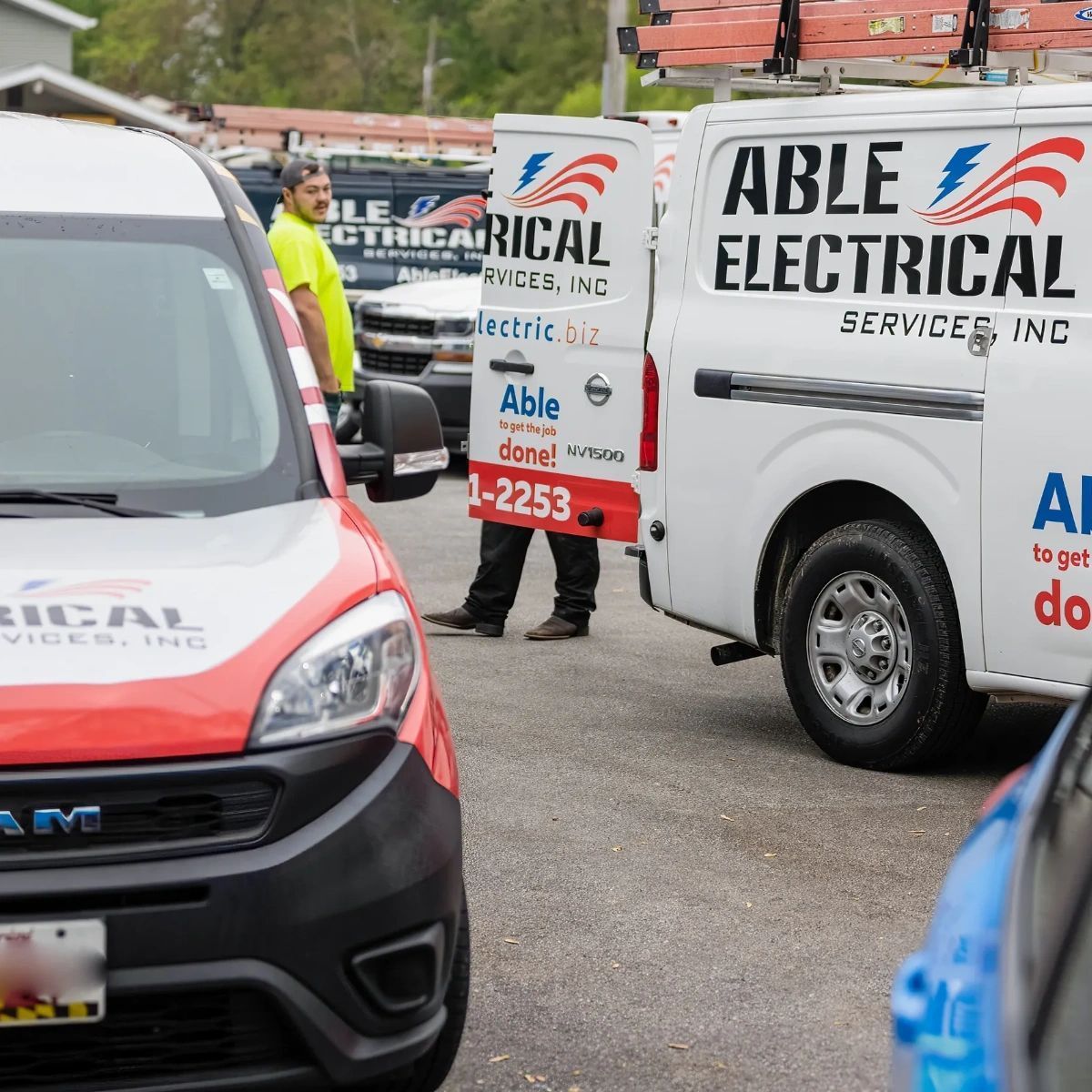 A person in a yellow safety shirt stands by an Able Electrical Service van with an open door in an outdoor parking lot.
