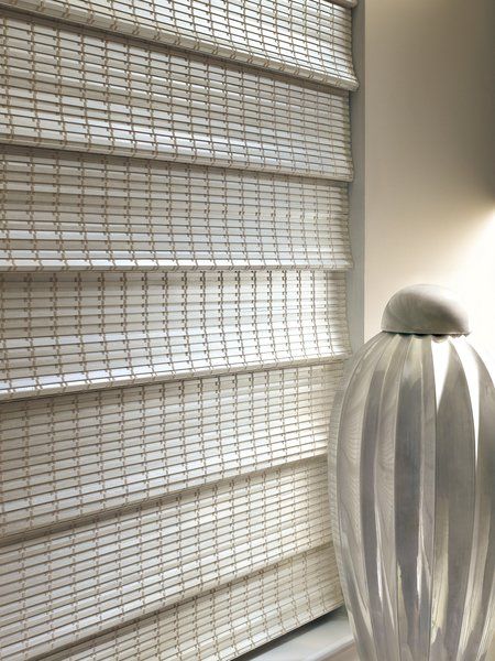 A white vase is sitting in front of a window with blinds.