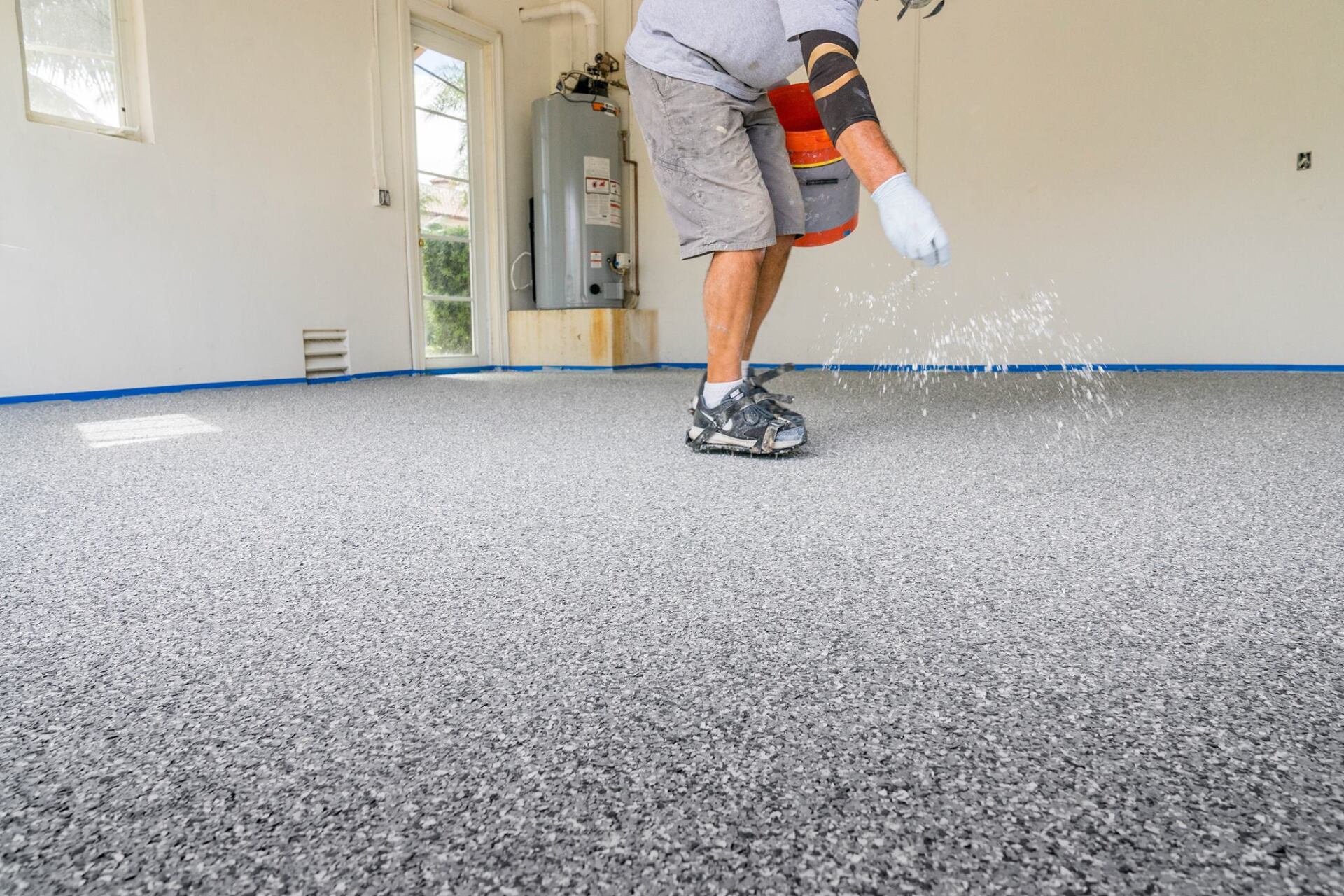 Person applying gray flakes to a freshly coated epoxy garage floor, indoors.