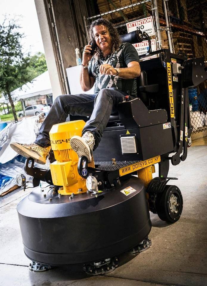 Man on a yellow and black industrial floor grinder, talking on the phone in a warehouse.