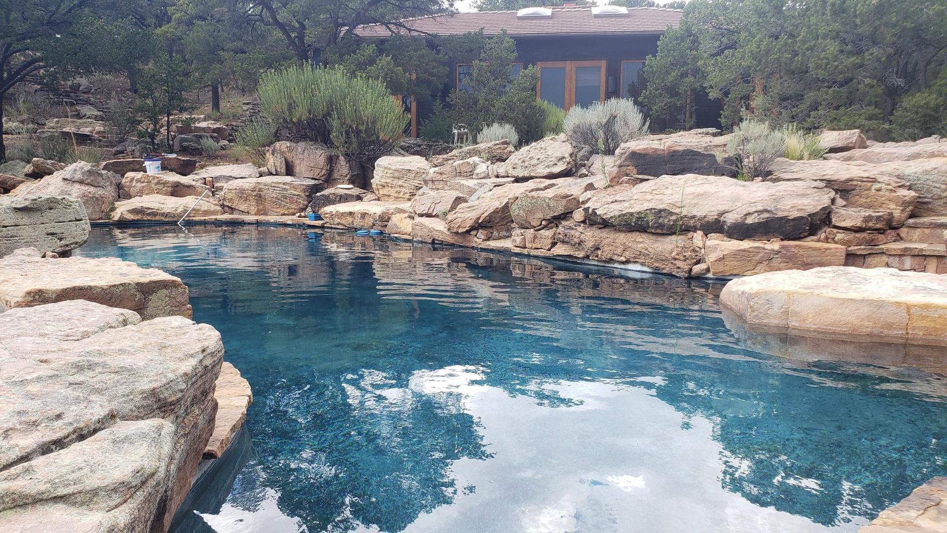 A Large Swimming Pool Surrounded by Rocks and Trees With a House in the Background