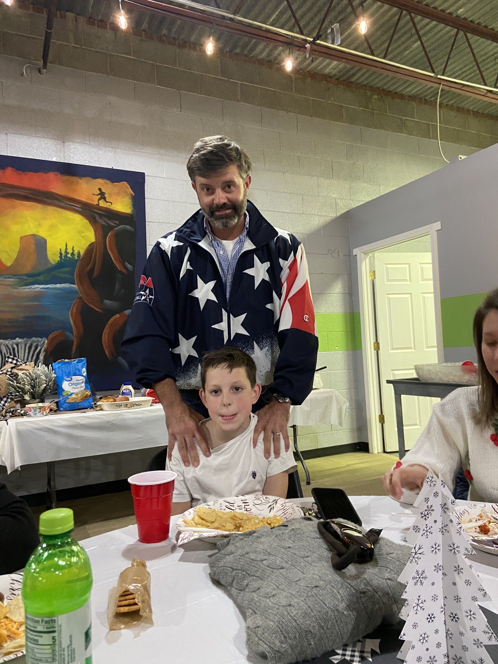 Man in star-patterned jacket with a boy at a table, hands on the boy's shoulders. Party setting.