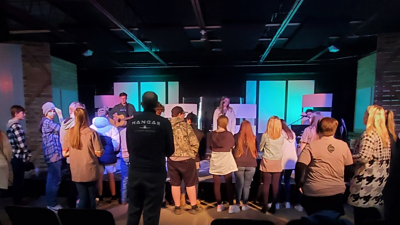 A group of people, mostly teenagers, gather on a stage with a band, illuminated by stage lights. The setting appears to be a church or community center.