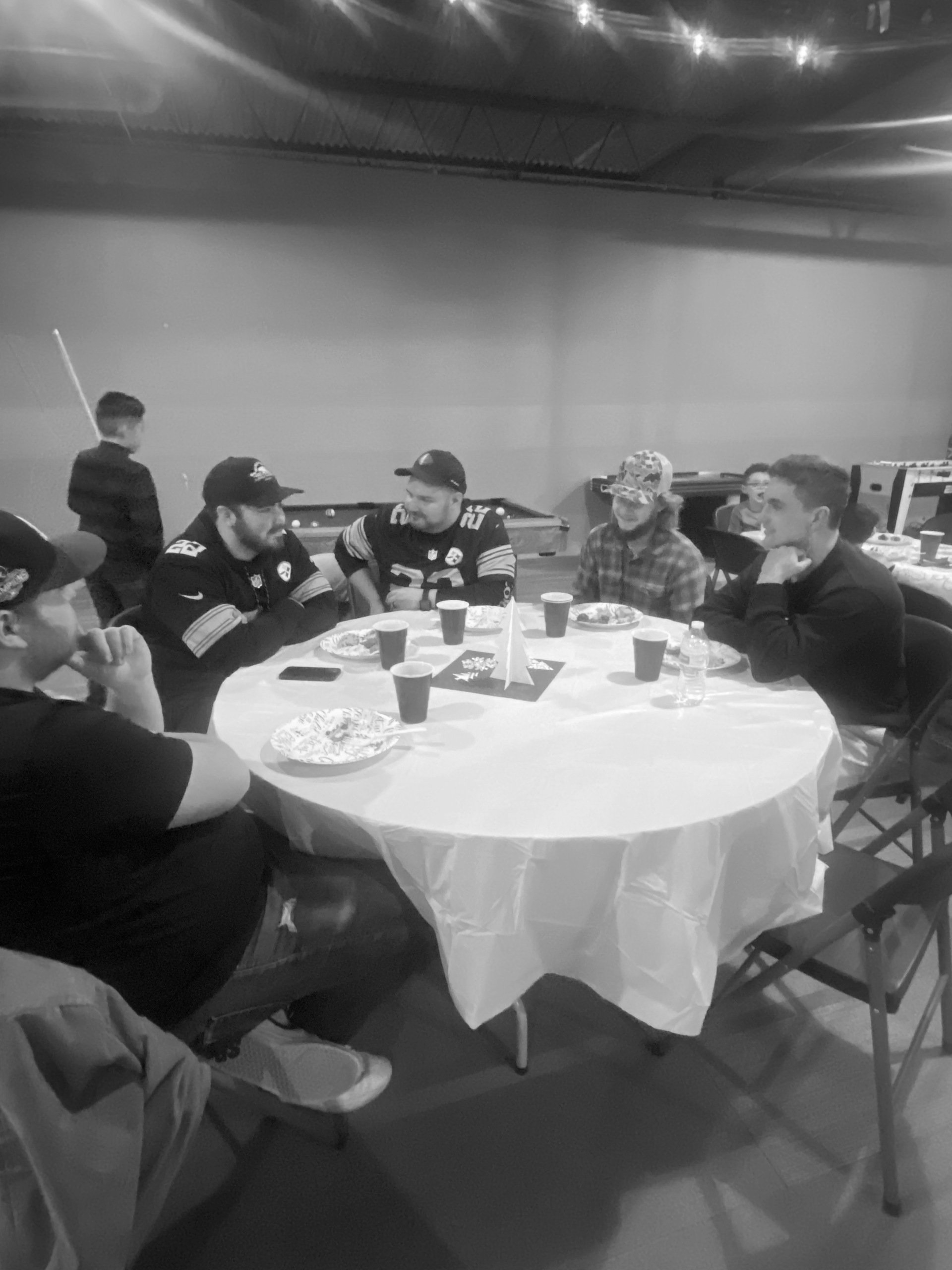 A group of six people sit around a round table with a white tablecloth, engaged in conversation. They are in an indoor setting, possibly a gathering.