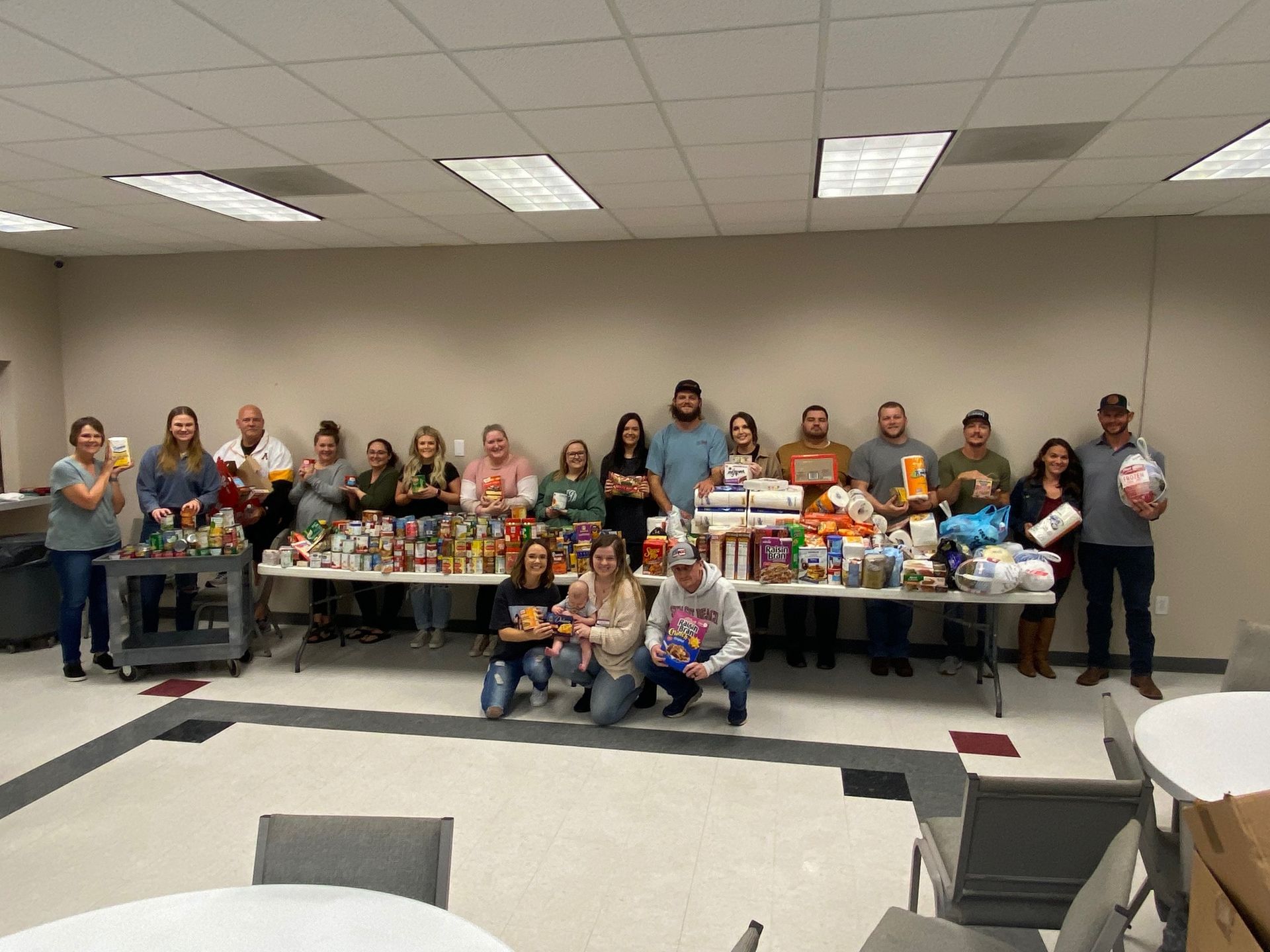 Group of people pose behind tables laden with donated food items in a brightly lit room, likely a community food drive.