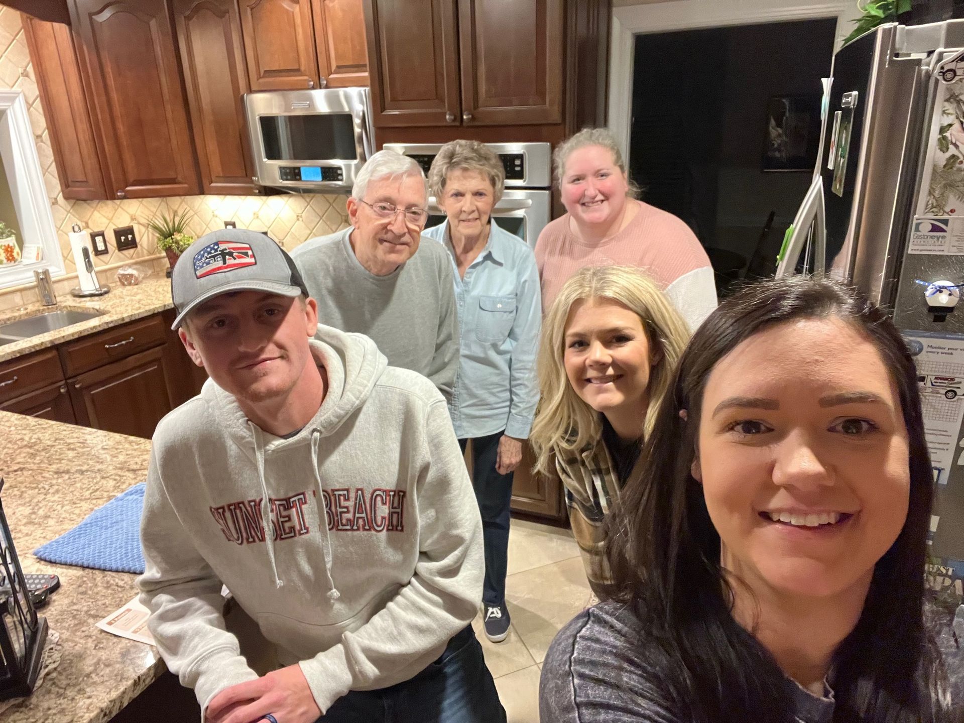 A group of six people smiling in a kitchen, with a woman taking a selfie. The kitchen has dark cabinets and stainless steel appliances.