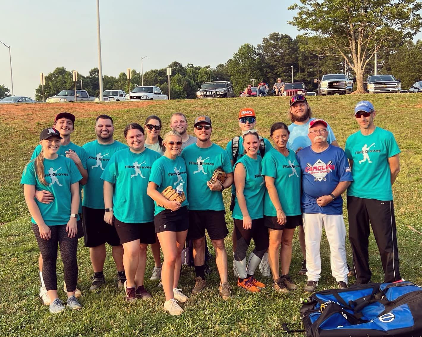 A softball team poses for a group photo on a grassy field. They wear matching teal shirts and black shorts, smiling for the camera.