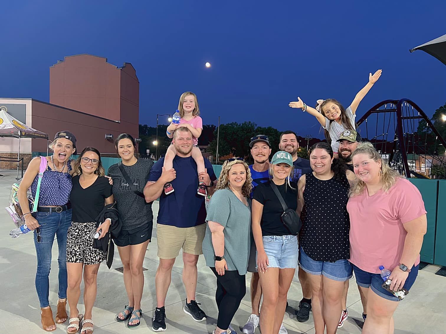 Group of people smiling and posing outdoors at dusk, with a child on shoulders and arms raised.