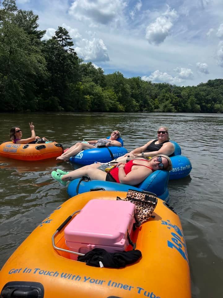 People floating on tubes down a river on a sunny day.  One person waves, while others relax with sunglasses, surrounded by lush greenery.