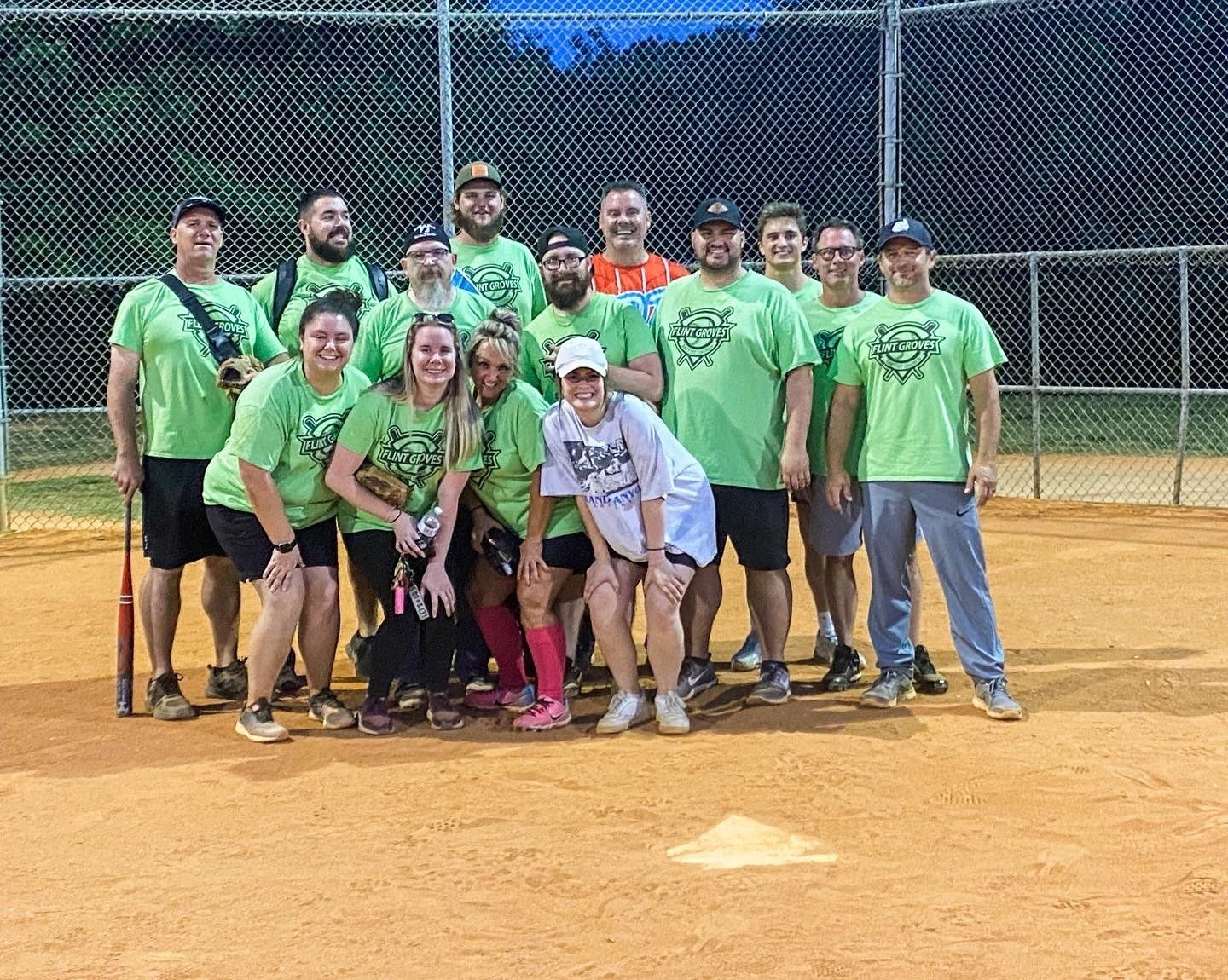 A softball team in green shirts poses on a dirt field. They are smiling and looking at the camera. A chain-link fence is in the background.