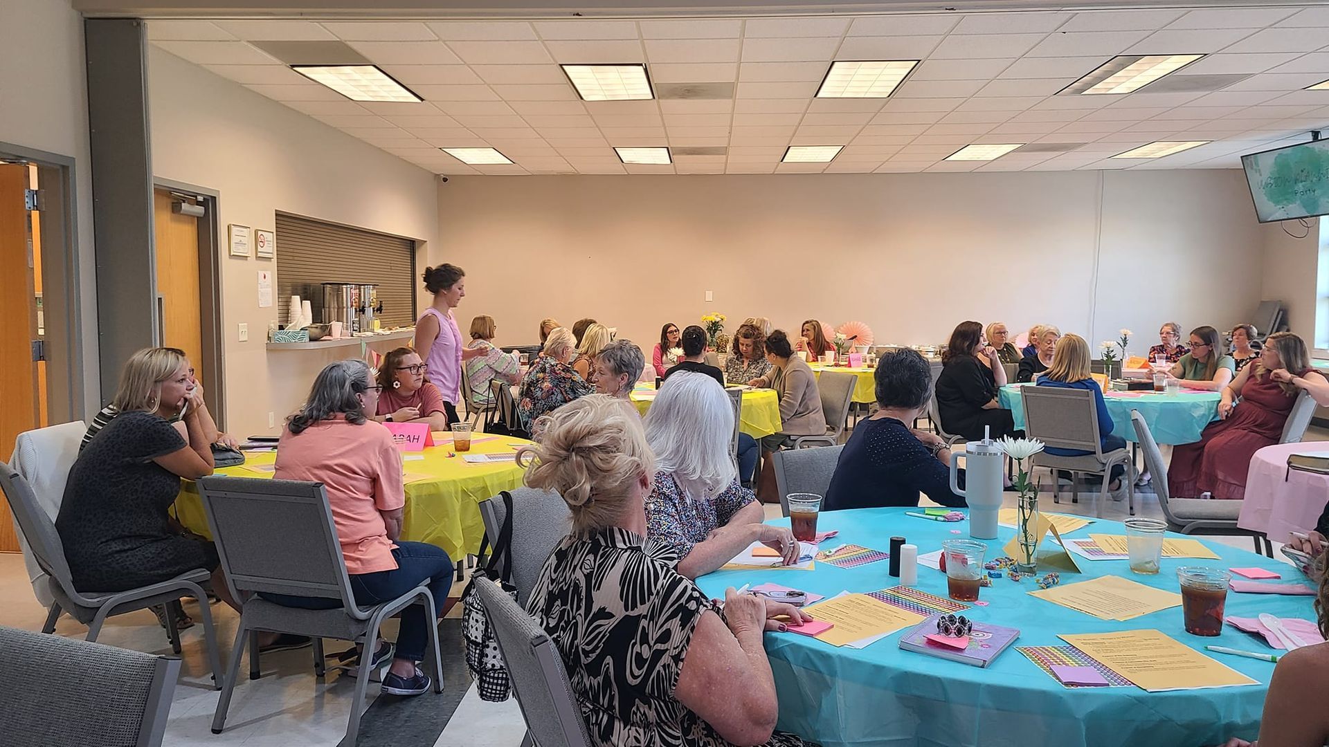 Women seated at tables, gathered in a brightly lit event space, some listening to a speaker. Tables are covered in colorful tablecloths.