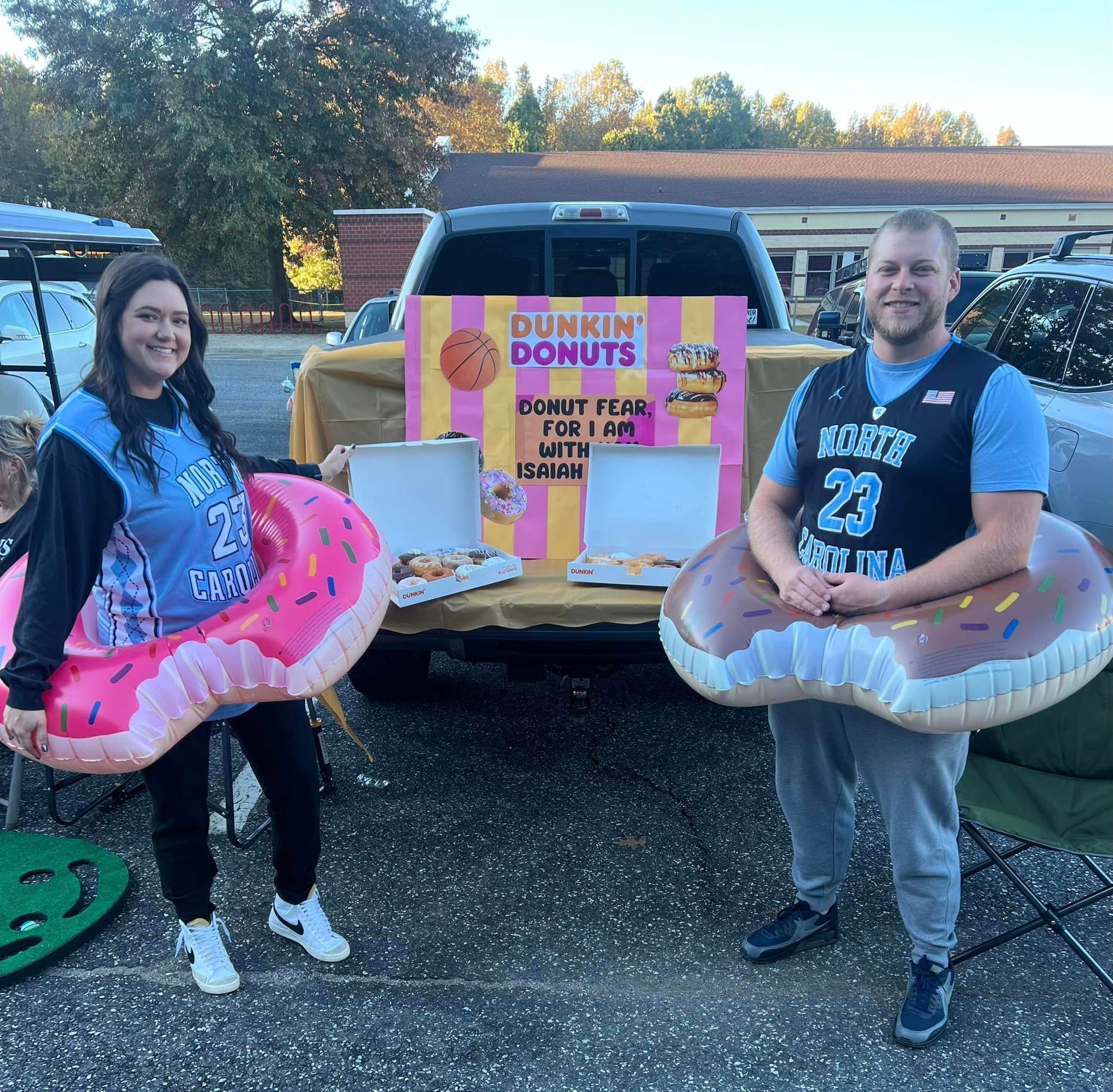 Two people in donut costumes, smiling, standing in front of a truck bed with Dunkin' Donuts sign and donuts.