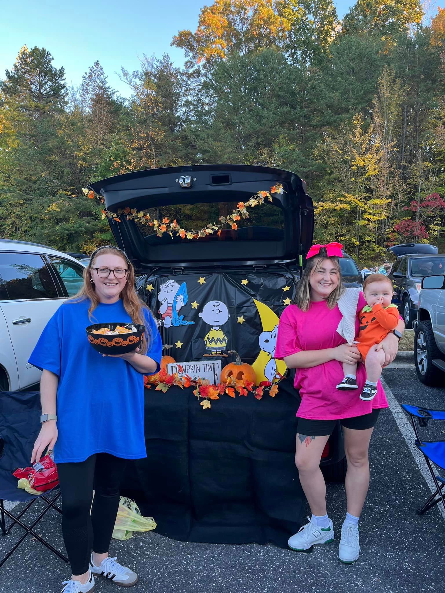 Two women and a baby dressed as Peanuts characters pose in front of a themed car trunk decorated for Halloween.