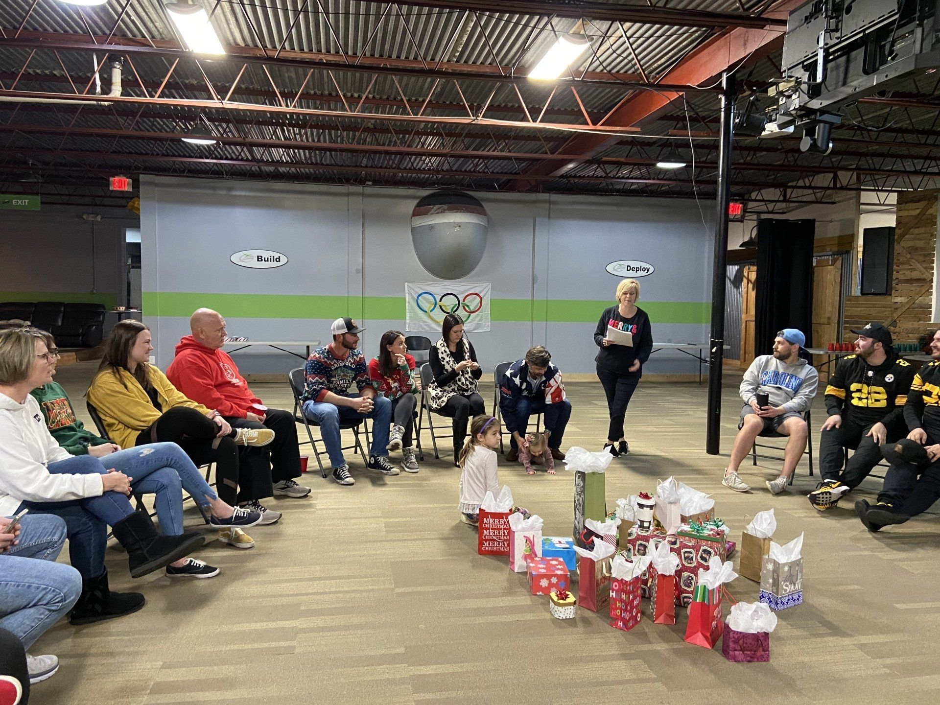 People gathered in a community center with gift bags. A woman speaks, while others watch. Decorations and an Olympic logo are on the wall.