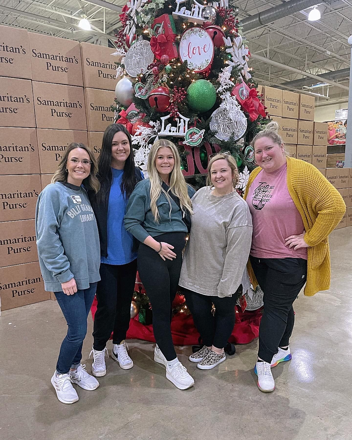 Five women pose in front of a decorated Christmas tree, smiling in a warehouse setting with boxes.