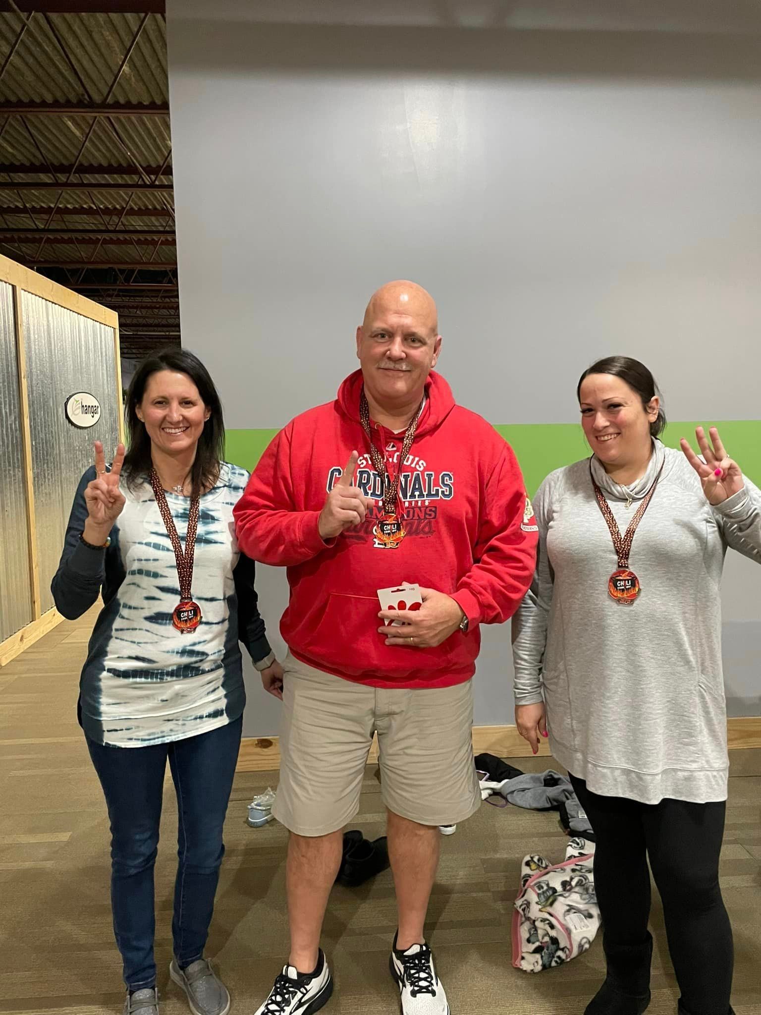Three people wearing medals pose in front of a wall. The woman on the left holds up a peace sign, the man in the middle holds up a thumbs-up and the woman on the right holds up three fingers.