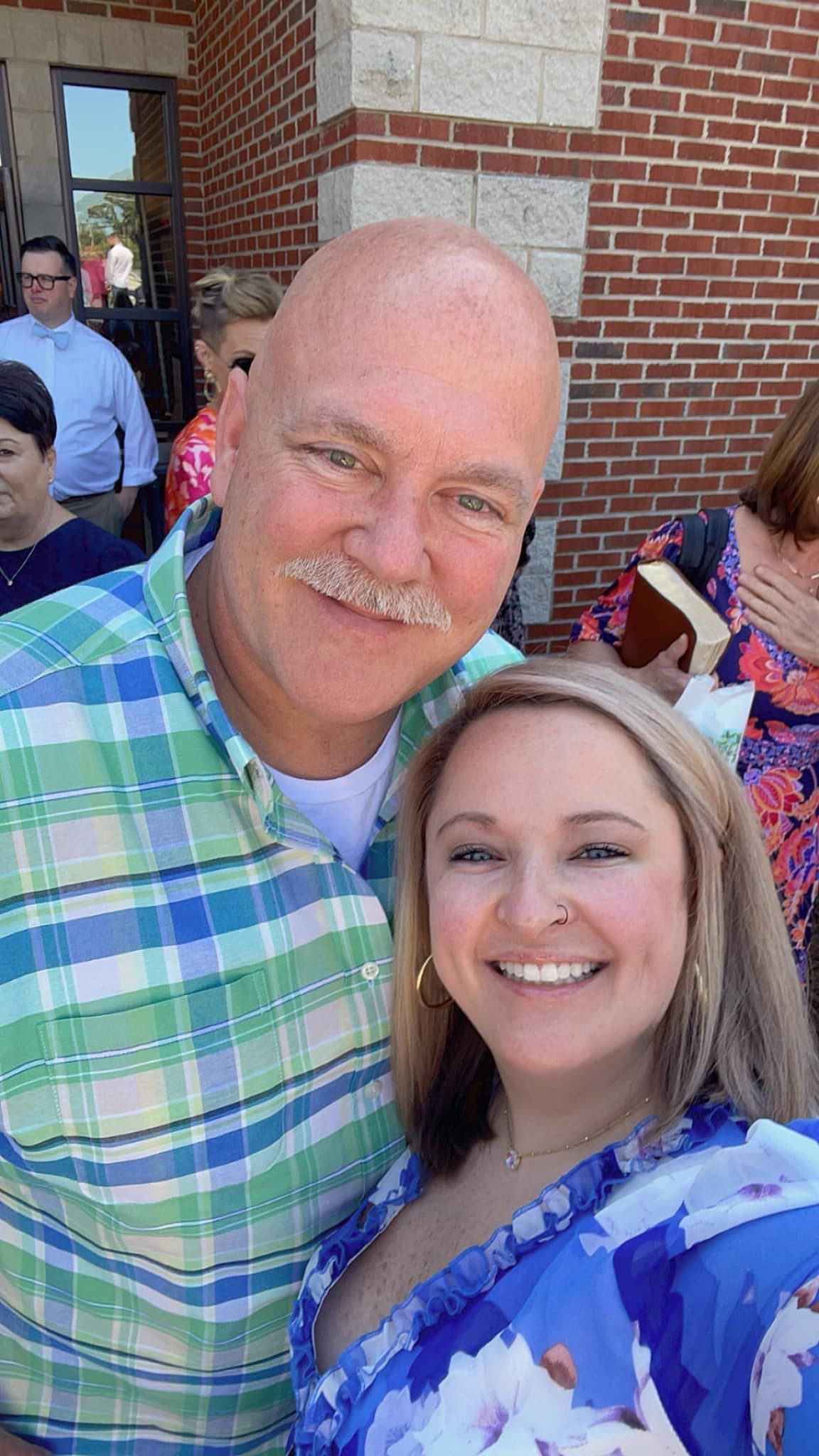A smiling man and woman take a selfie outdoors. The man has a mustache, wearing a blue and green plaid shirt. The woman has blonde hair and wears a blue floral dress.