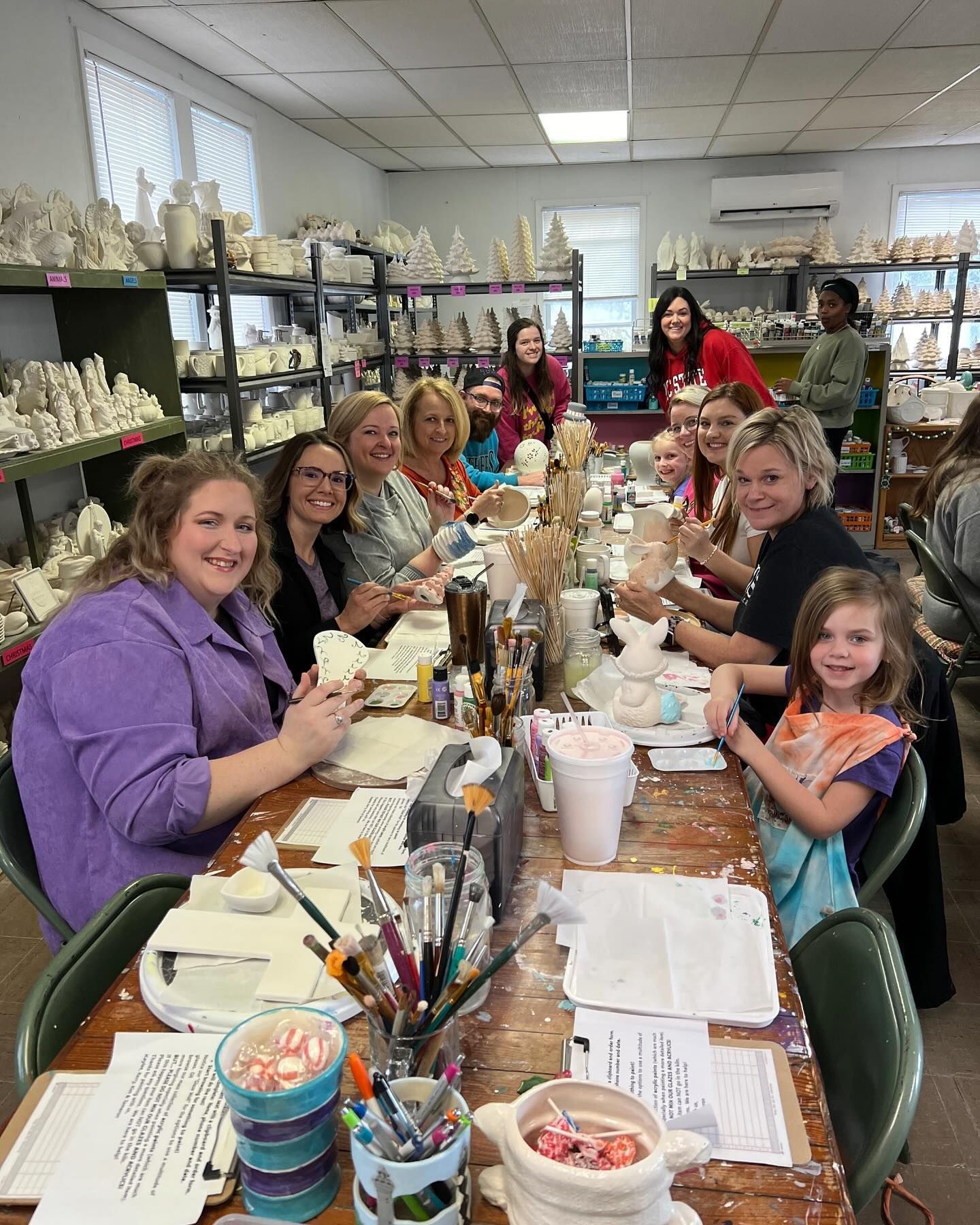 People painting pottery at a long table in a studio. Smiling women and a child are working on ceramics, surrounded by shelves of finished pieces.