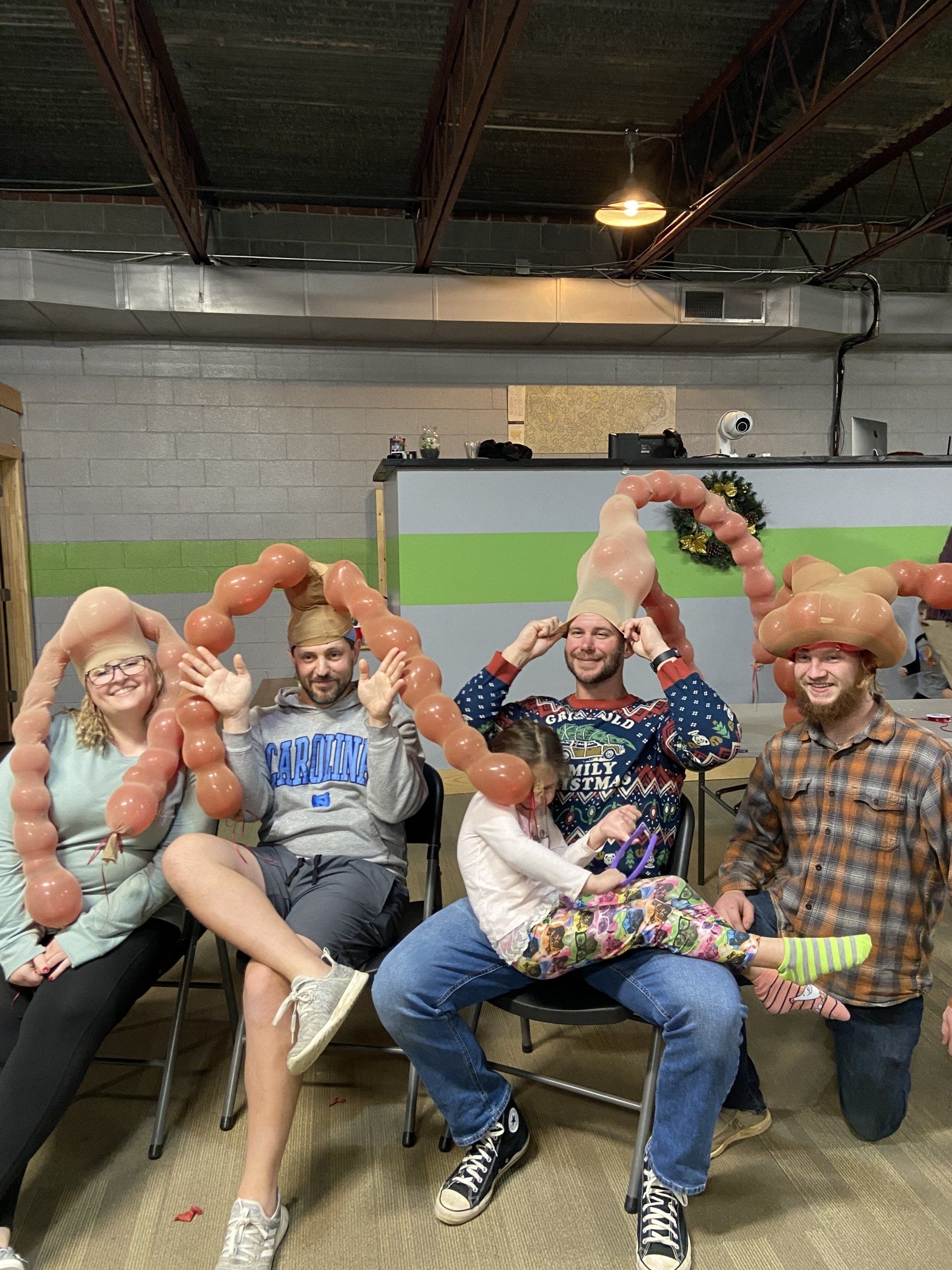 Five people sitting and kneeling indoors wearing brown balloon hats, some waving.