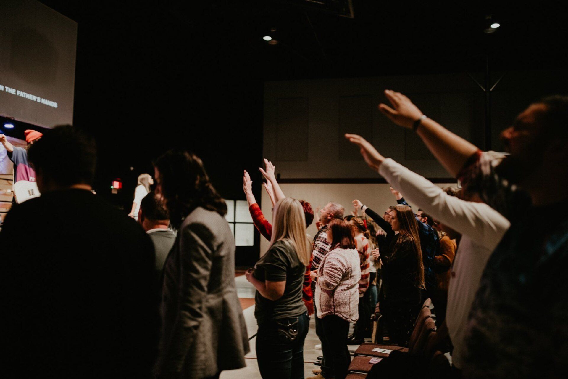 People in a dark auditorium raise their arms in worship. A stage and screen are in the background.