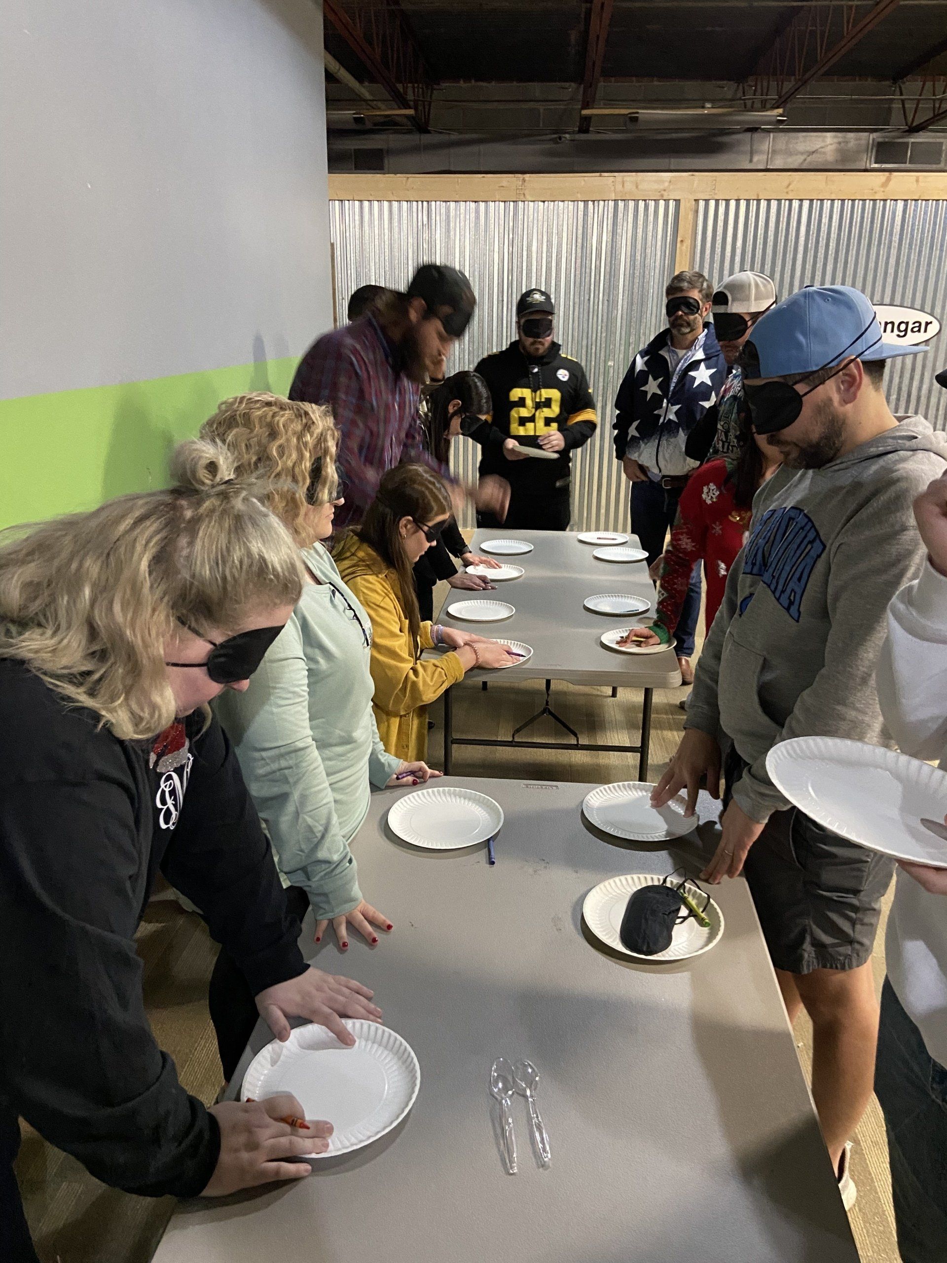 People with blindfolds at a table, likely tasting food. Plates, forks, and a casual setting suggest a game or activity.