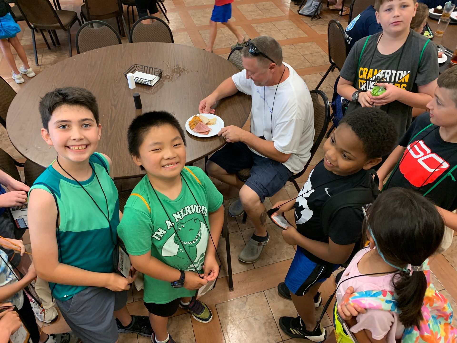 Group of children smiling at a table where a man is eating. Children are wearing colorful shirts, in a brightly lit room.