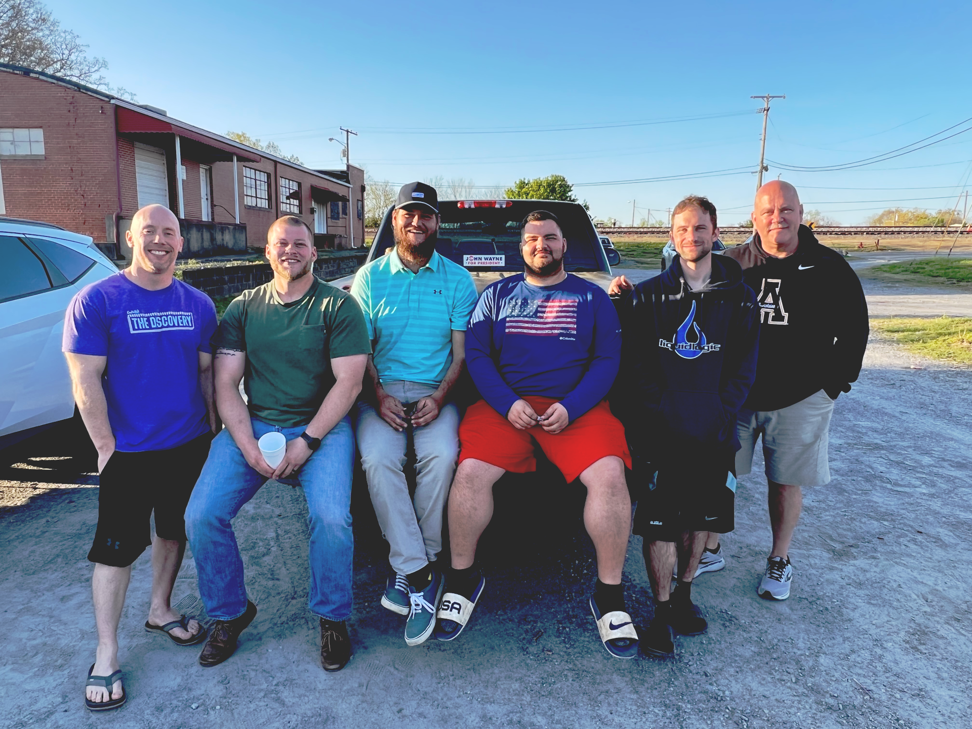 Six men pose on the back of a dark car outdoors. They are smiling, some wearing casual clothes, and near a brick building on a sunny day.