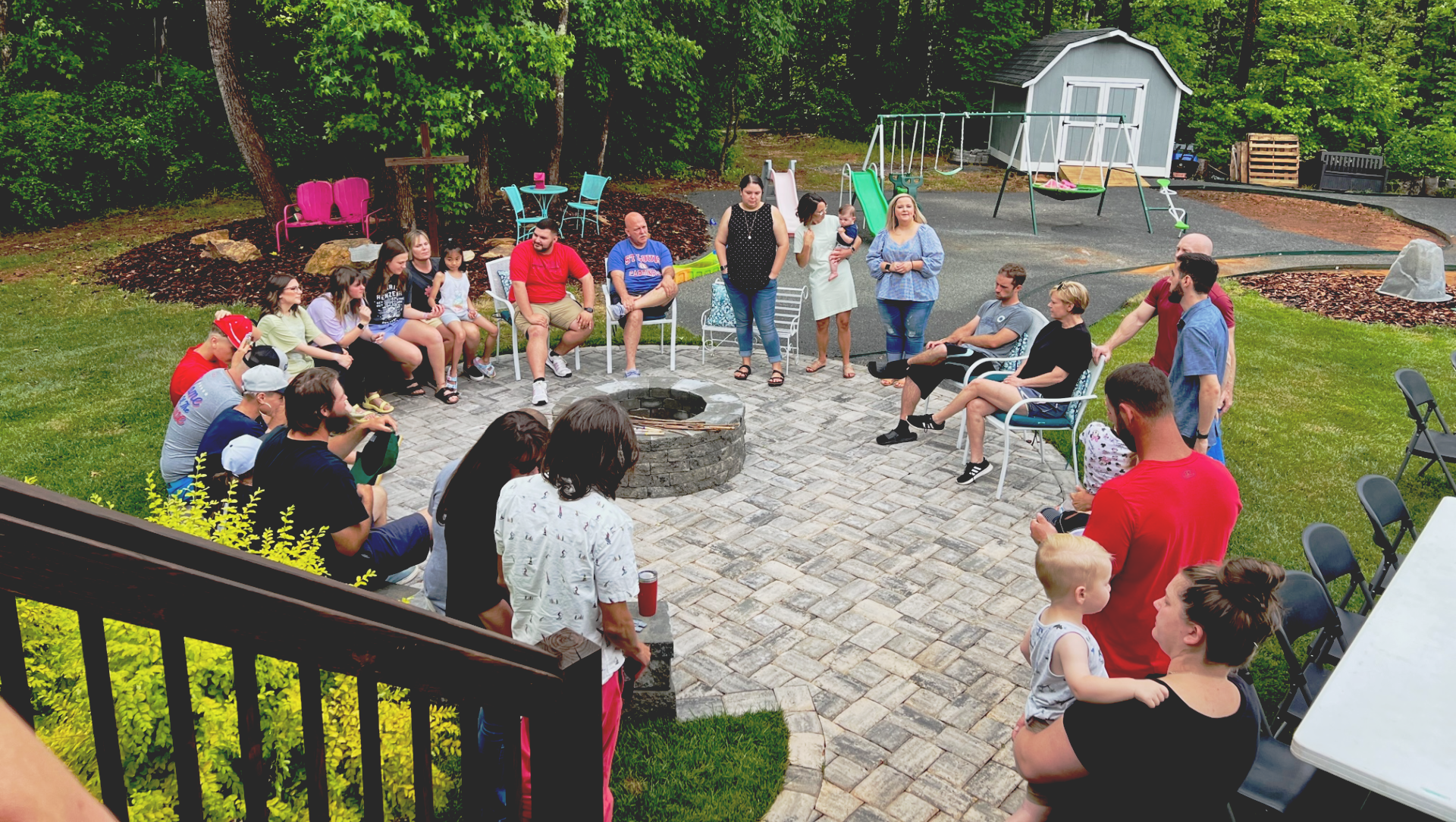Group of people gathered outdoors around a fire pit. People sit, stand, and converse on a brick patio.