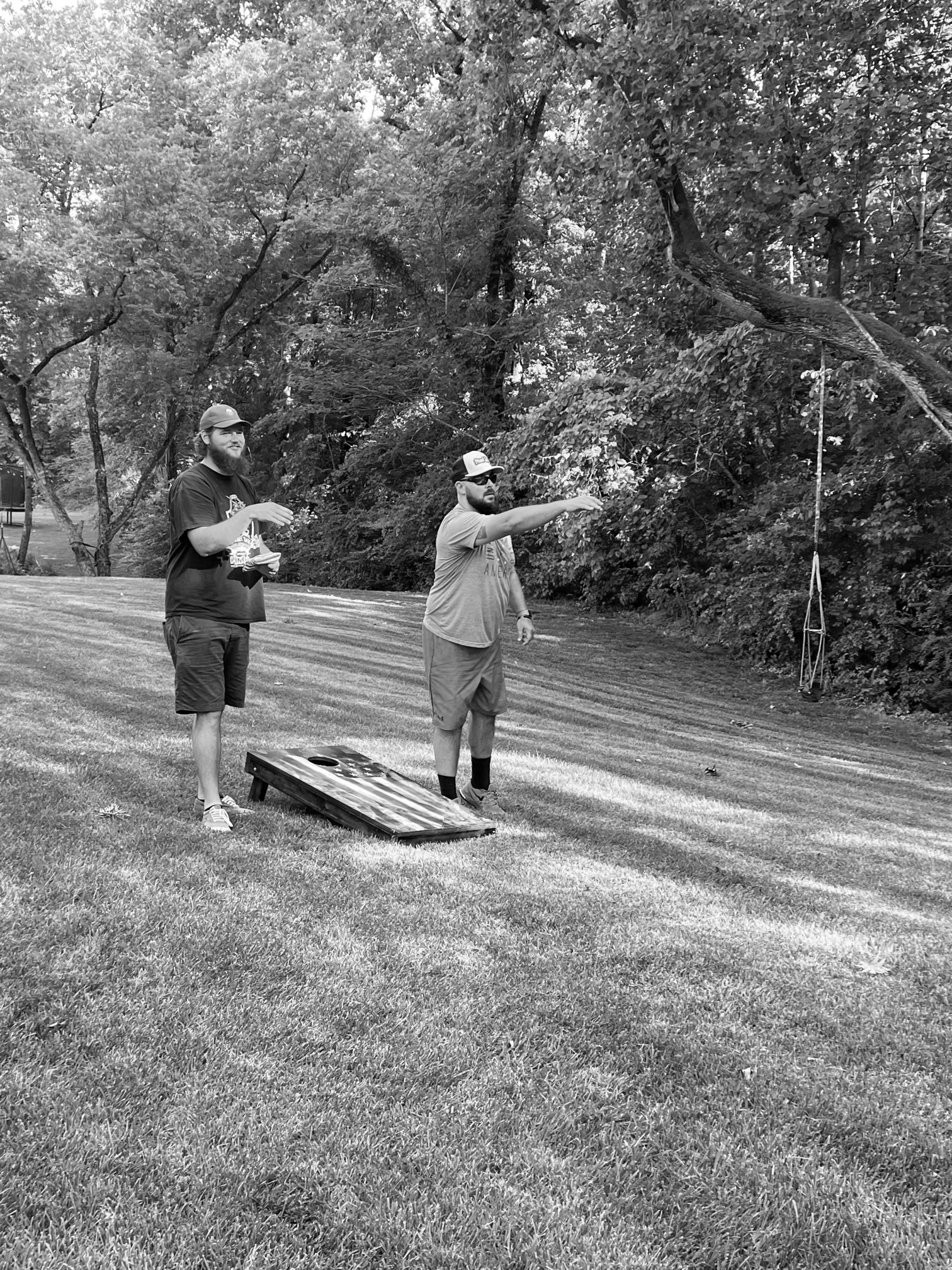 Two men playing cornhole on a grassy lawn in a park. One throws a beanbag; the other watches.