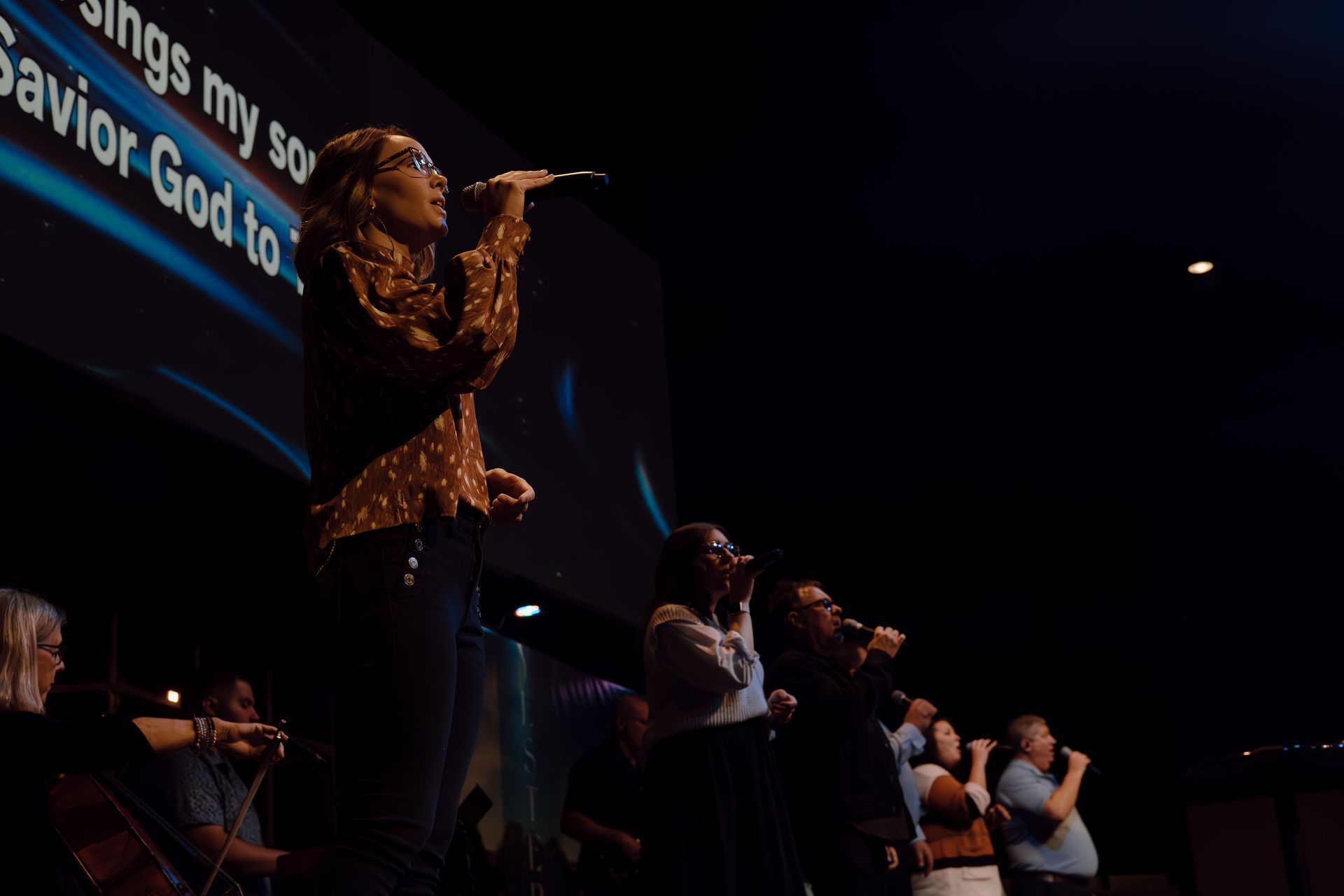 A woman singing on stage with a microphone, leading a choir in a dark auditorium. The lyrics of a hymn are on a screen behind them.