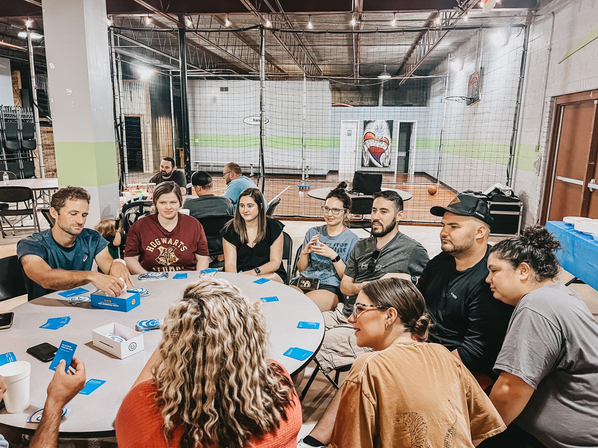 A diverse group of people sits around a table, playing cards in an industrial-looking space. They appear to be engaged in conversation and enjoying themselves.