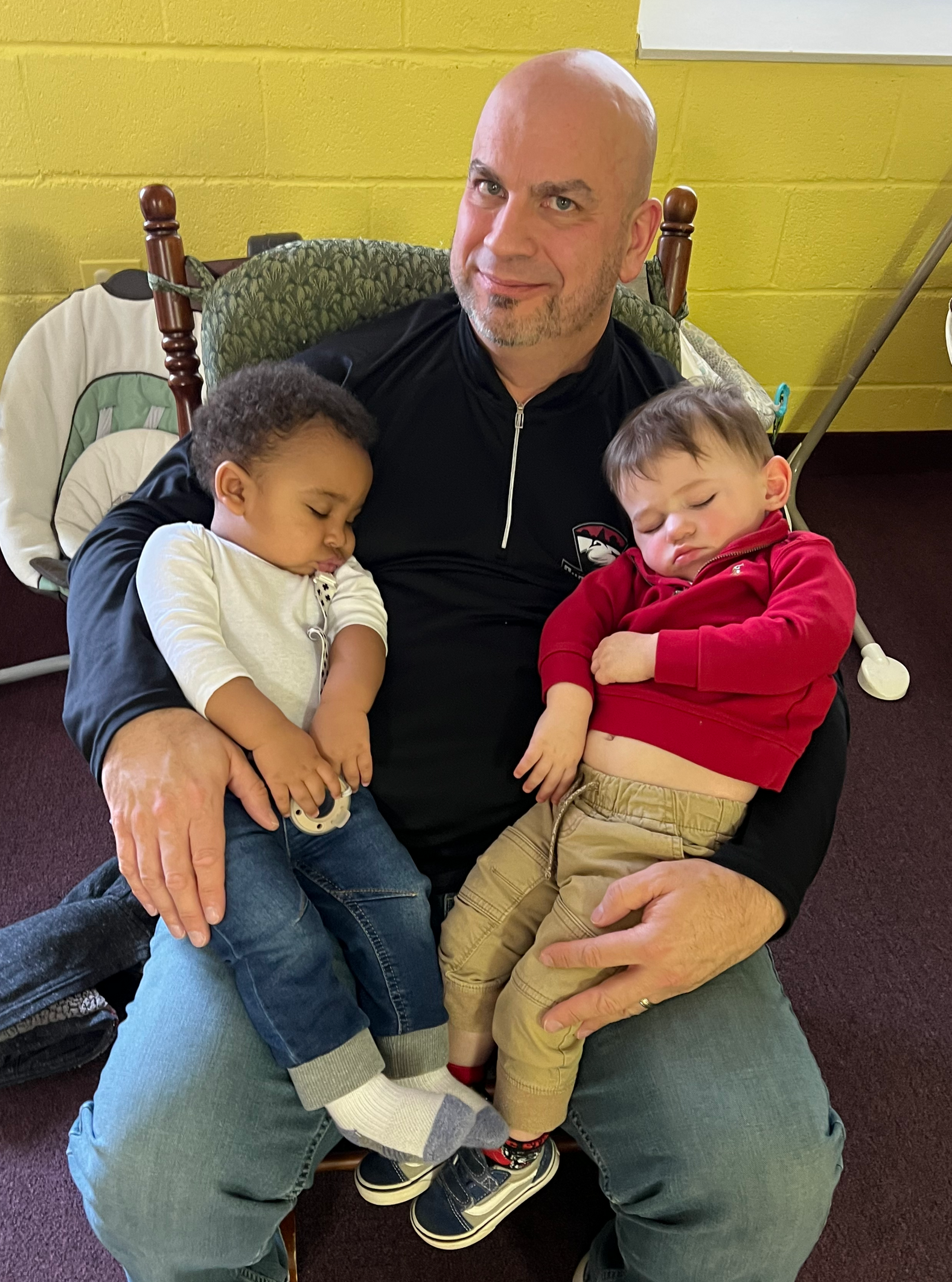 A man sits holding two sleeping babies, one black and one white, in his lap. They are indoors, and he's smiling.