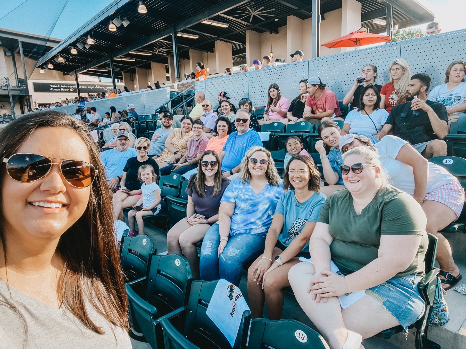 Group of people smiling at a baseball game. A woman in sunglasses takes a selfie in the foreground; many people in the stands behind her.