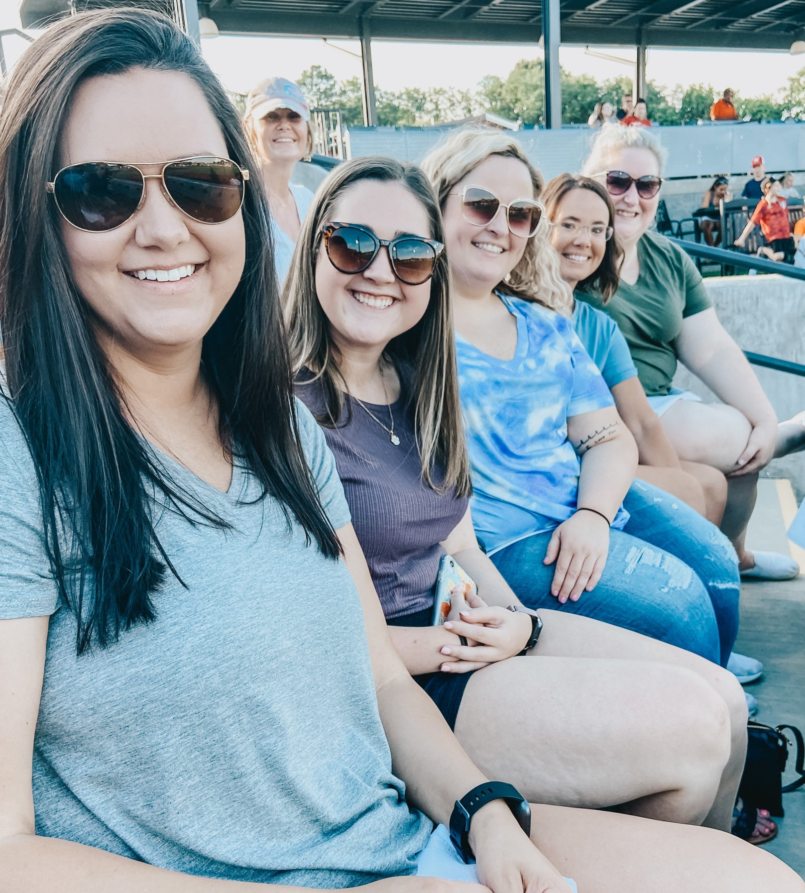 Group of smiling women wearing sunglasses, sitting closely together outdoors in a stadium or similar setting.