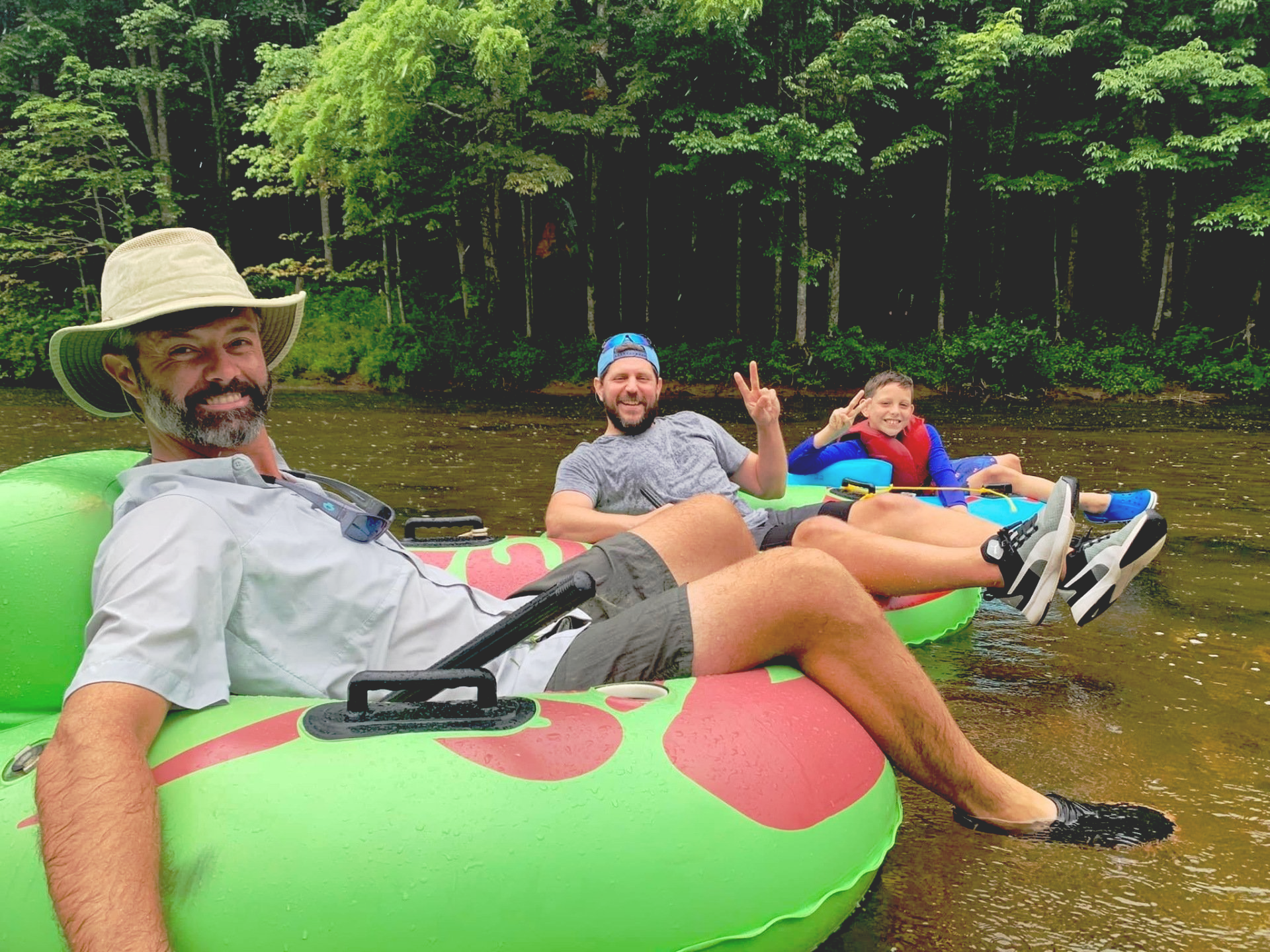 Three people float down a river in green inner tubes. A man in a hat smiles, another man gives a peace sign, and a child smiles. Trees line the riverbank.