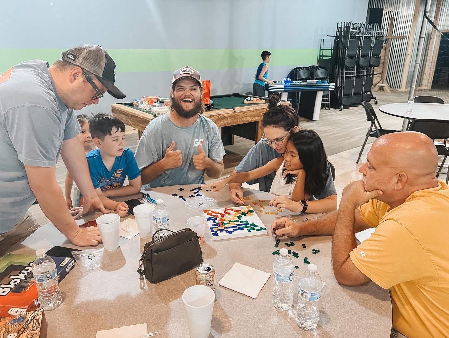 A group of people are gathered around a table playing a board game. They are smiling and appear to be enjoying themselves in a brightly lit room.