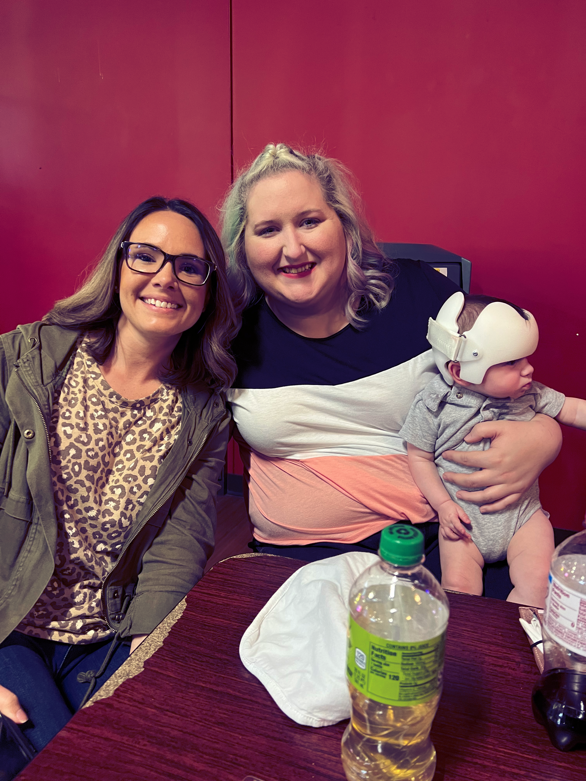 Two women and a baby smiling at the camera. The baby wears a helmet. They are seated indoors by a red wall.