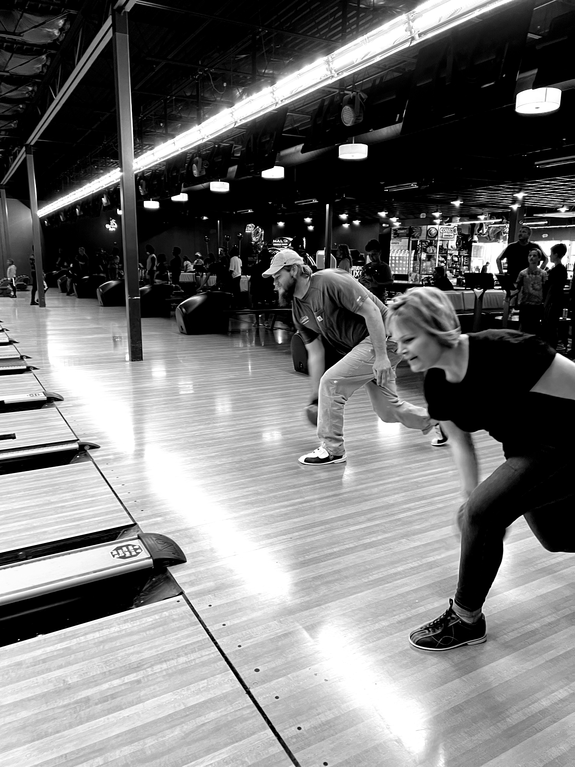 Two people bowling in a dimly lit bowling alley. The man is throwing the ball, and the woman is following through.