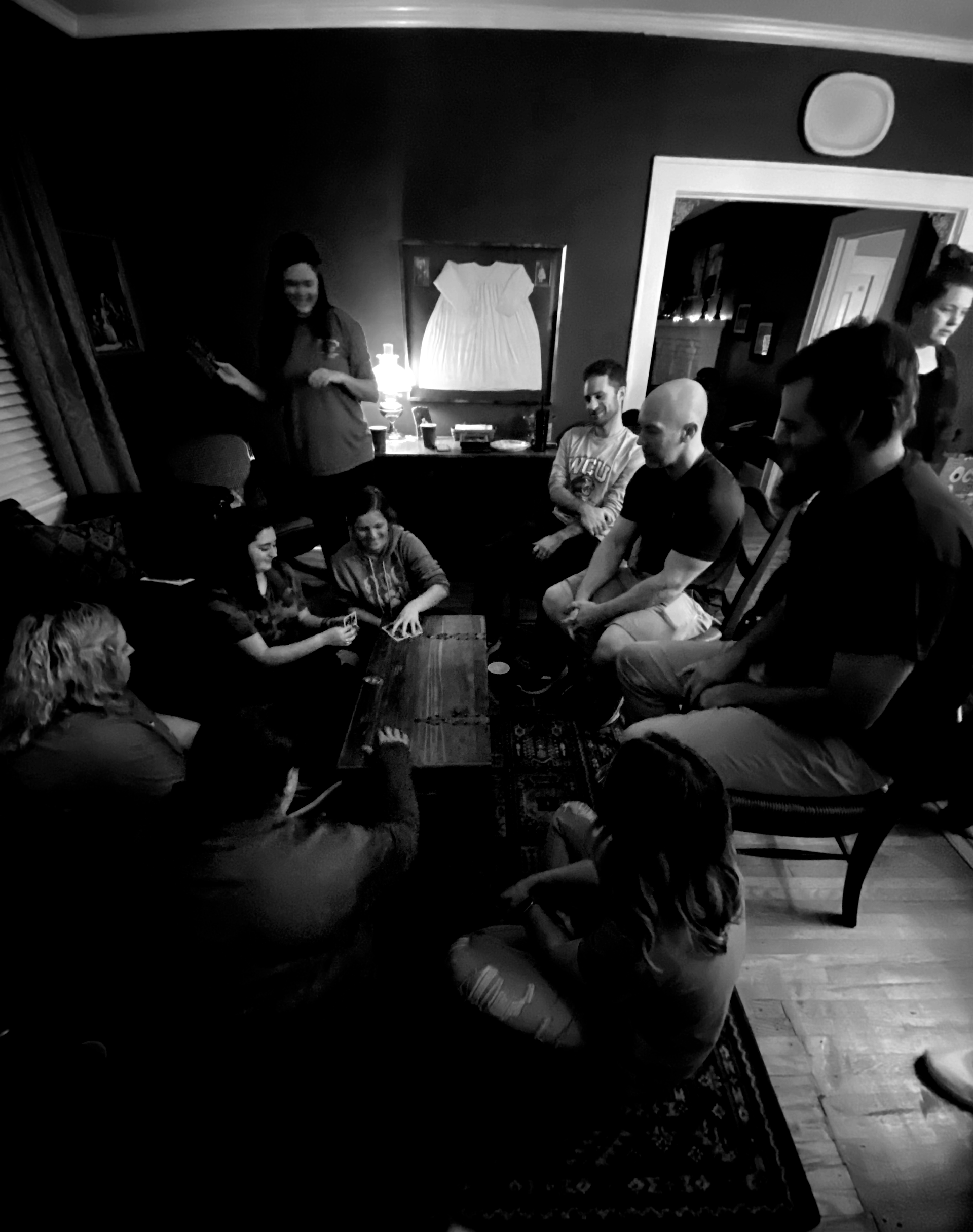 People gathered in a dimly lit room, sitting in a circle. Some are looking at each other, while one woman stands and gestures.