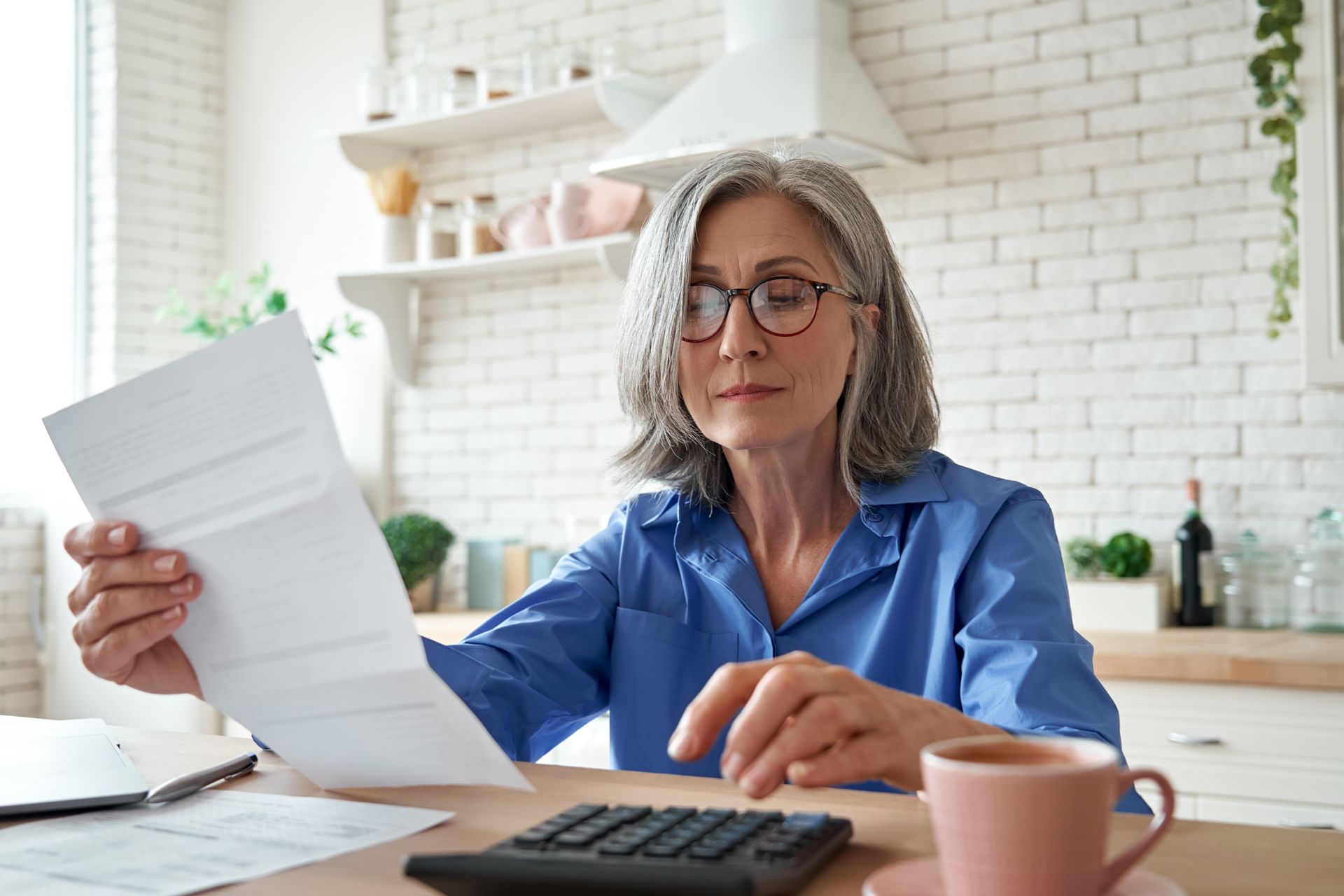 An older woman is sitting at a table reading a letter and using a calculator.