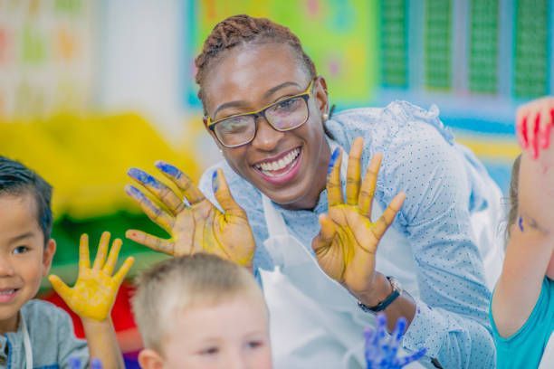 A teacher is standing next to a group of children with their hands painted yellow.