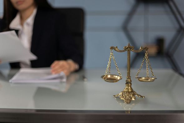 Golden scales of justice on a desk; a lawyer in a suit reviews documents in the background.