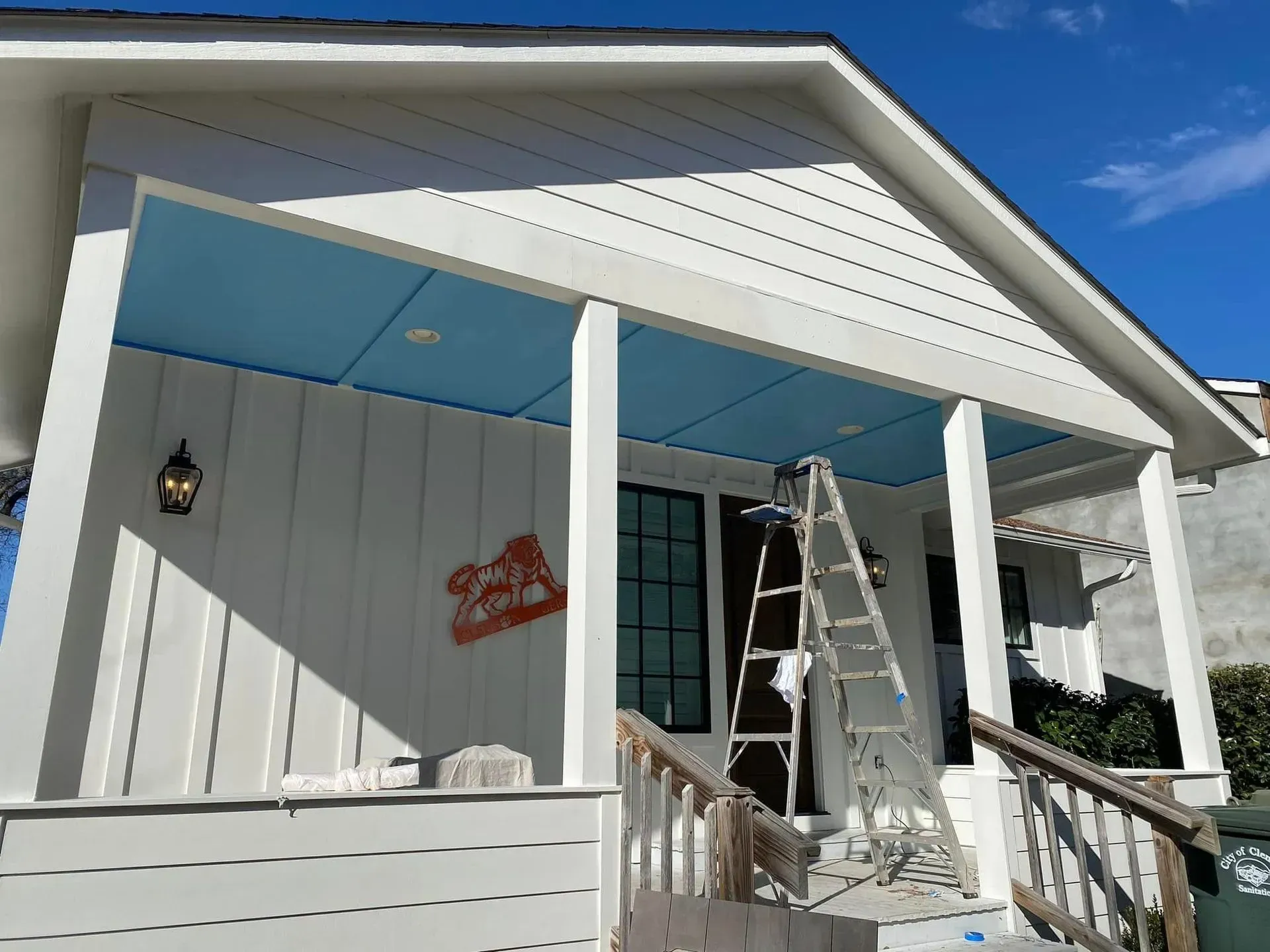 Front porch with white siding, blue awning, and a ladder beside the entrance under bright sunlight