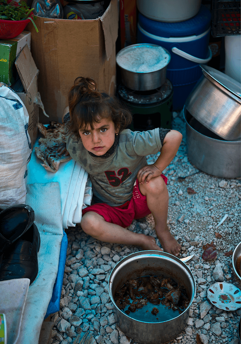 A yazidi child on sitting the floor among kitchen utensils and a food pan infant of her