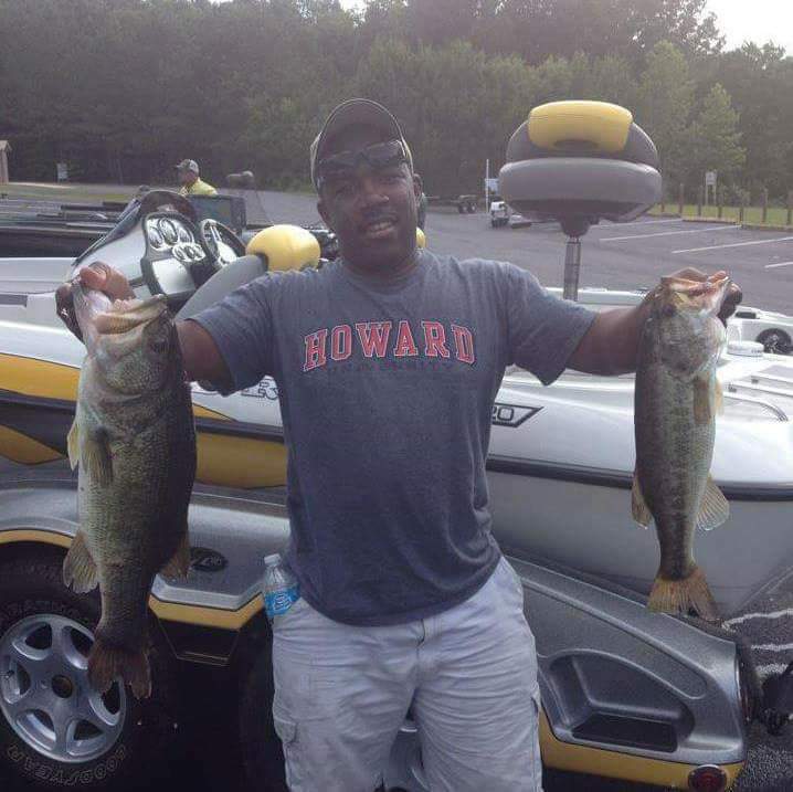Man holding two large bass by a boat, wearing a Howard University shirt, with a parking lot background.